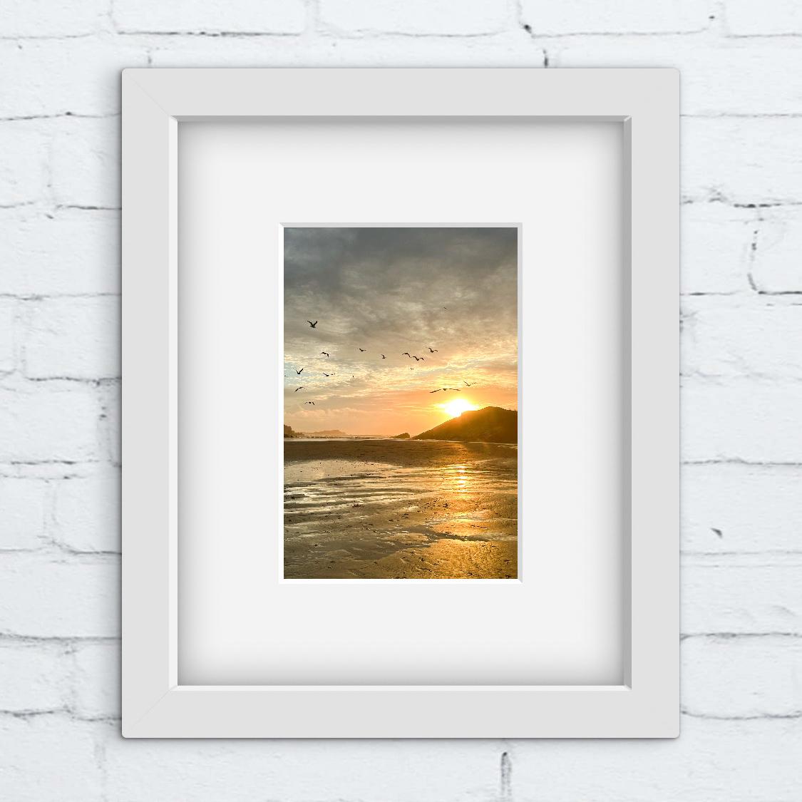 Framed artwork of a sunset over water with birds on a white brick wall.