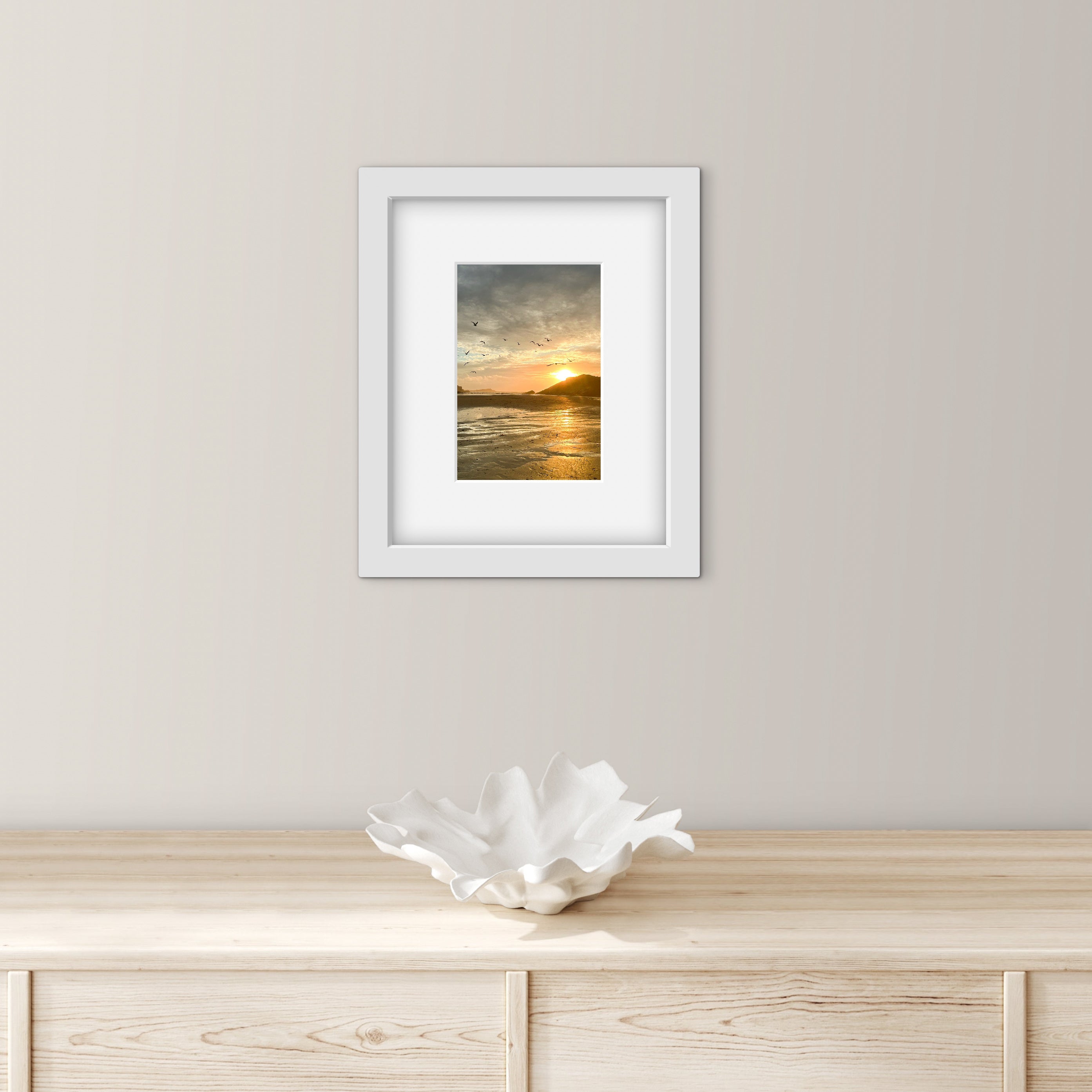 Framed artwork of a sunset over water on a wall above a wooden surface with a decorative bowl.