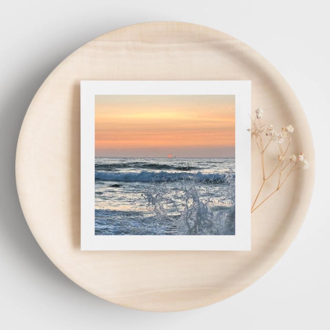 Greetings card of a sunset over the ocean with sea splash, displayed on a beige plate.