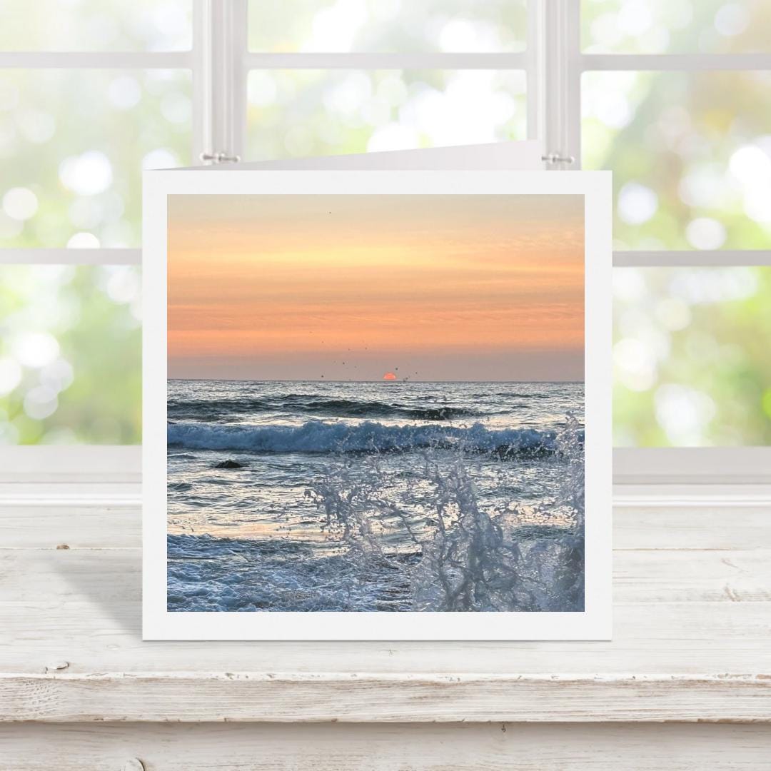 Greetings card of a sunset over the ocean with sea splash, displayed on a wooden surface with a blurred window background.