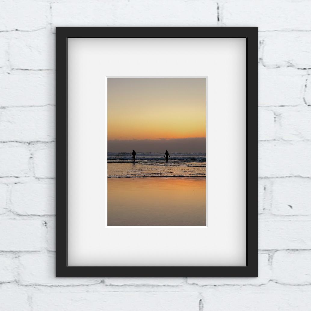 Framed photograph of two people going for a swim in the sea at sunset. Framed against a white brick wall