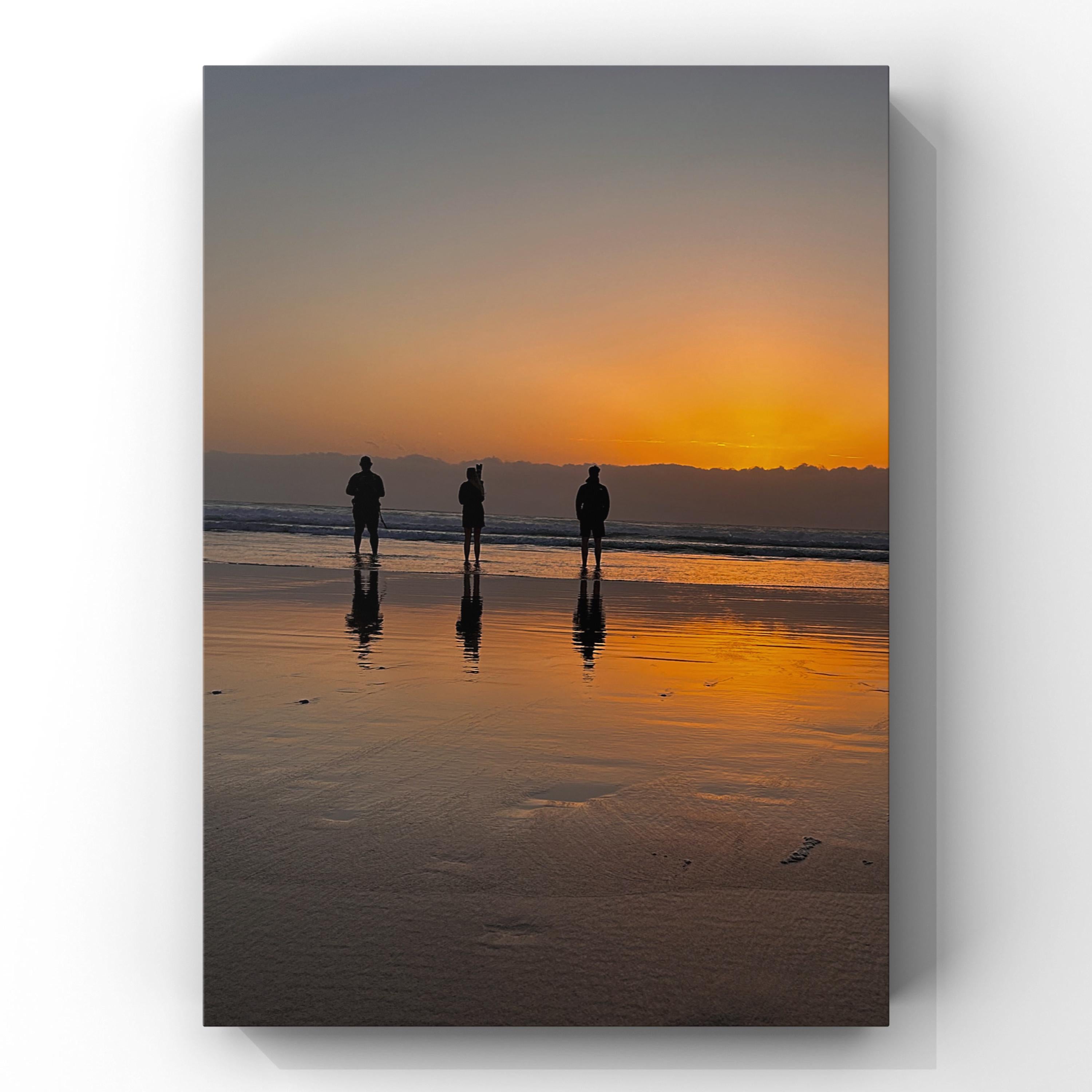 Three people standing on a beach at sunset, reflected in the wet sand.