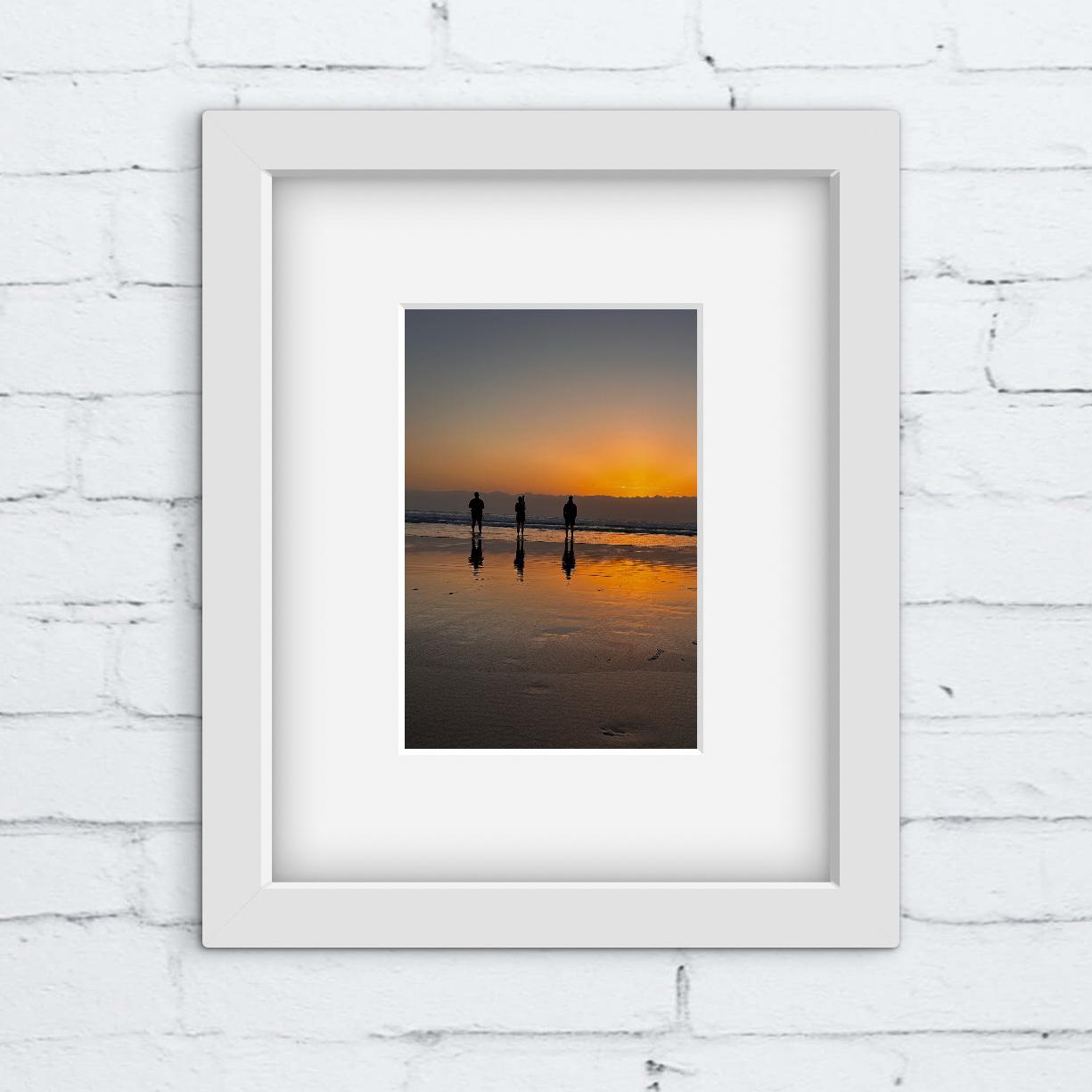 Framed photograph of a sunset on a beach with three people silhouetted against the water.