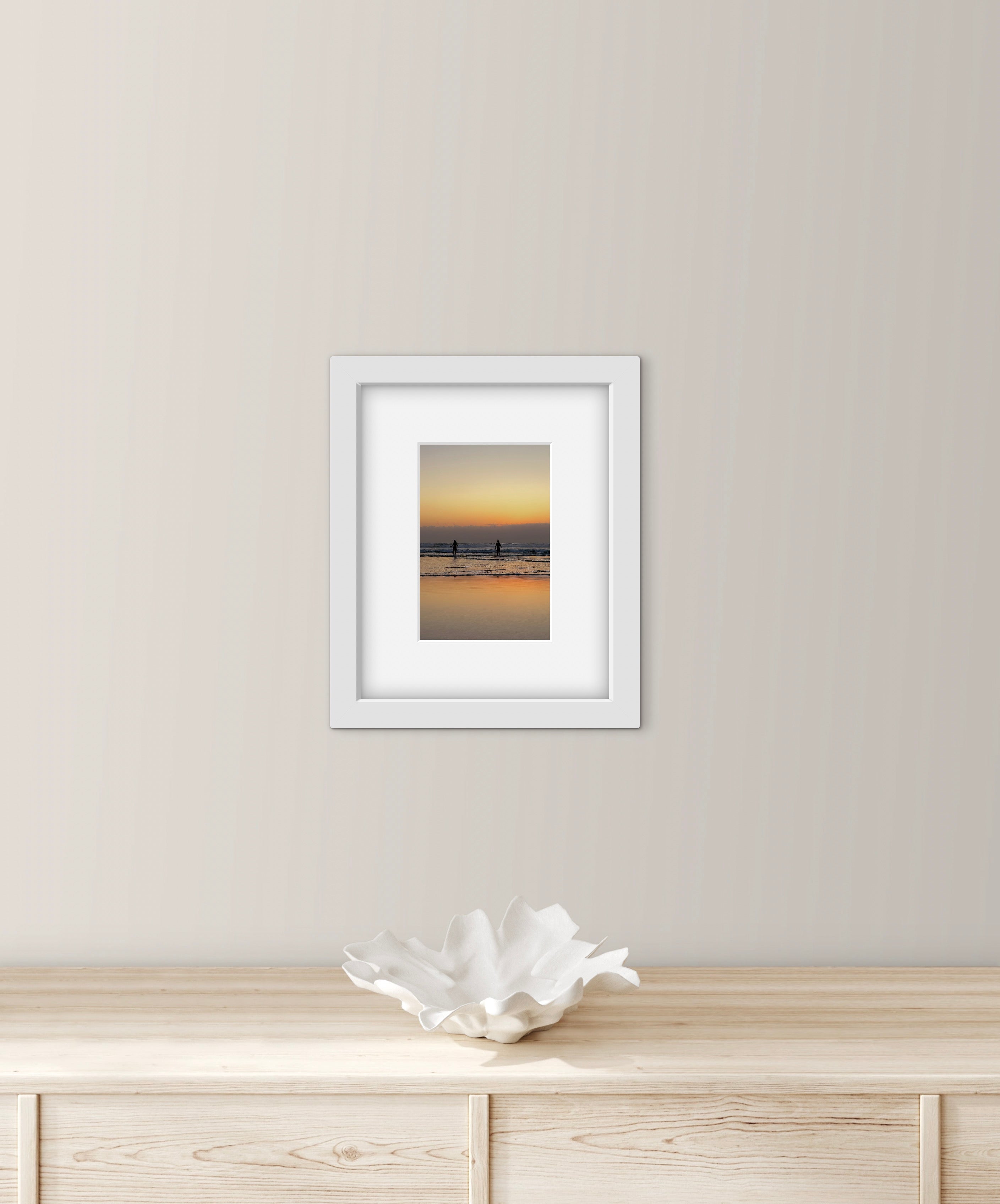 Framed artwork of two people going for a sea swim at sunset on a wall above a wooden surface with a decorative bowl.