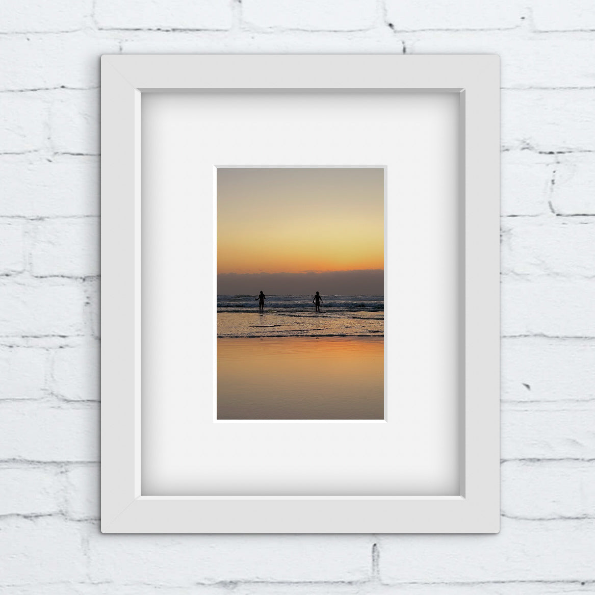 Framed artwork of a sunset over water with two silhouettes, on a white brick wall.