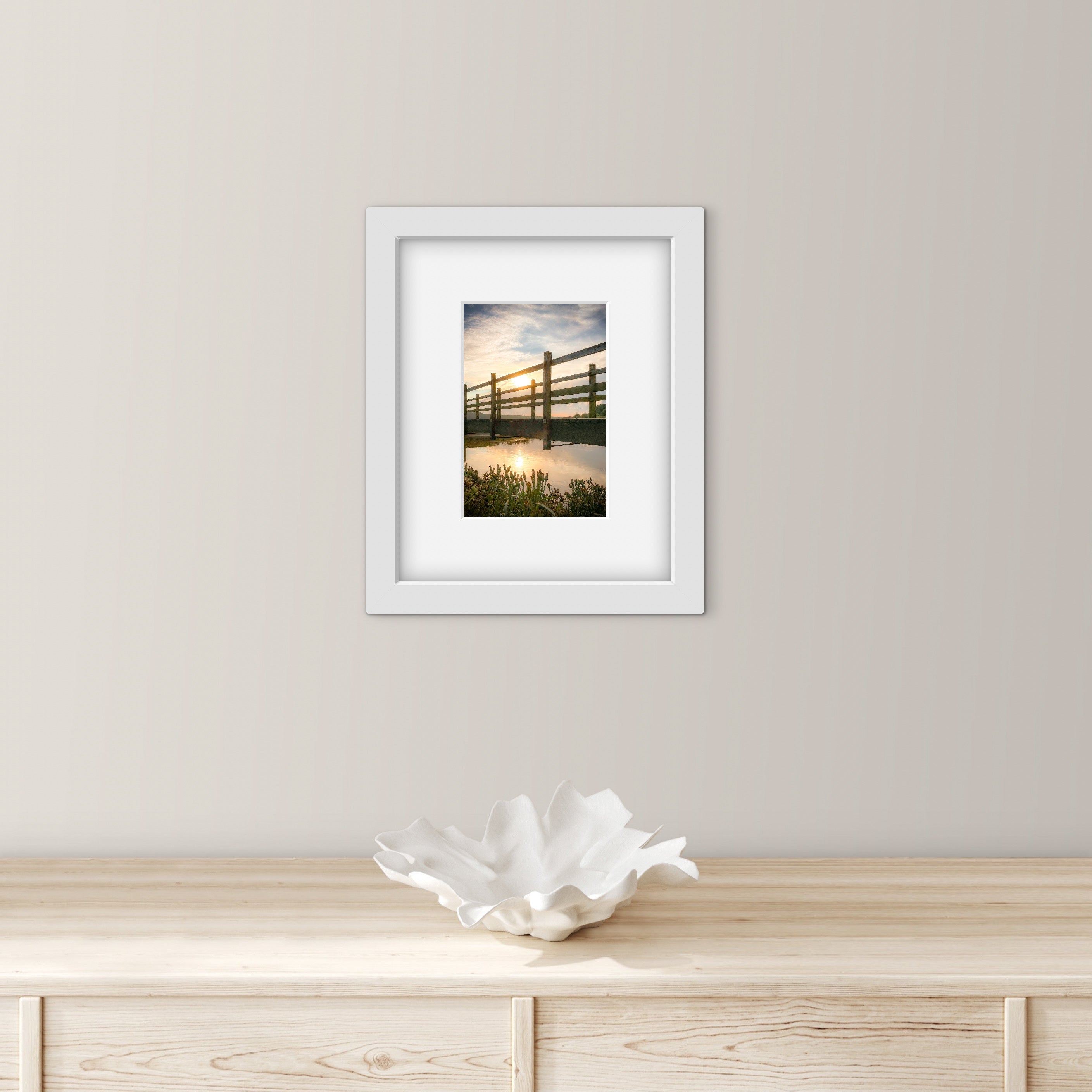 Framed photograph of a bridge over water  and sunset, on a wall above a wooden cabinet.