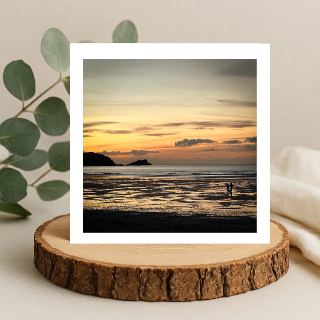 Sunset over a beach with two people standing on the shore, greetings card framed by a white border and sitting on a wooden coaster.