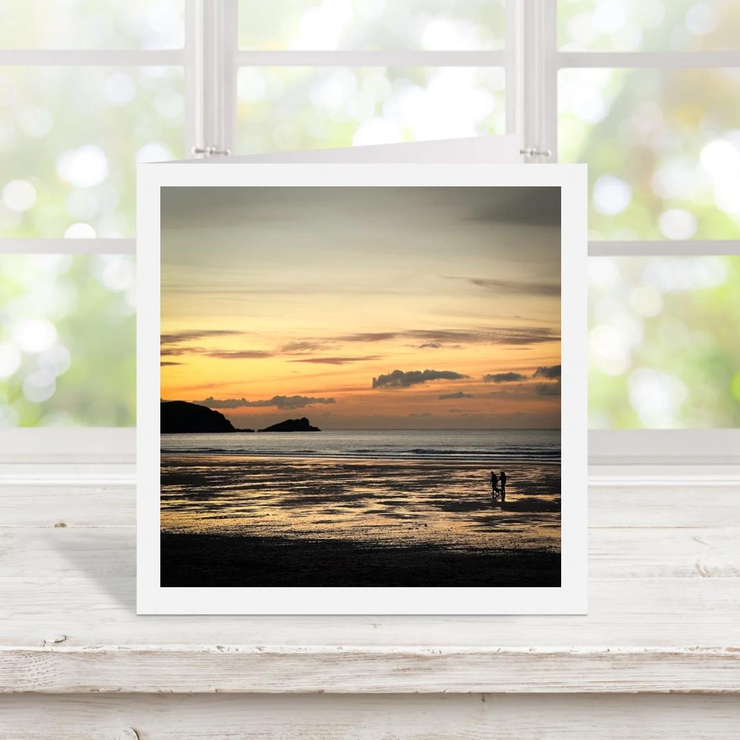 Square greetings card with photograph of a sunset over a beach with two people standing on the shore, displayed on a white surface.