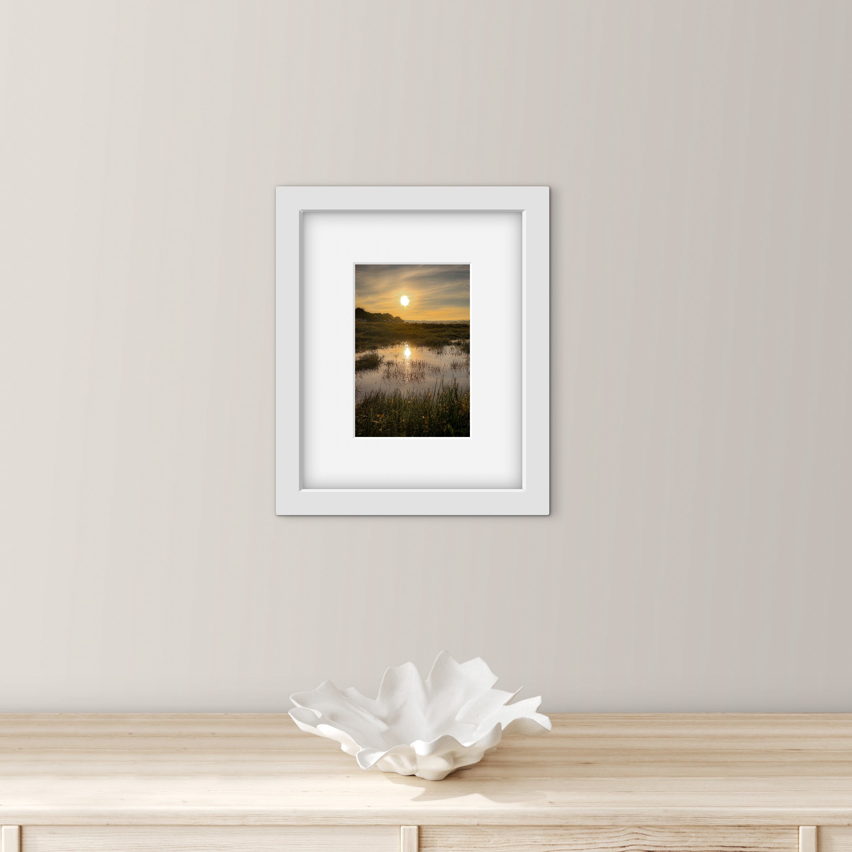 Framed artwork of a sunrise over marshy water on a wall above a wooden surface with a decorative bowl.