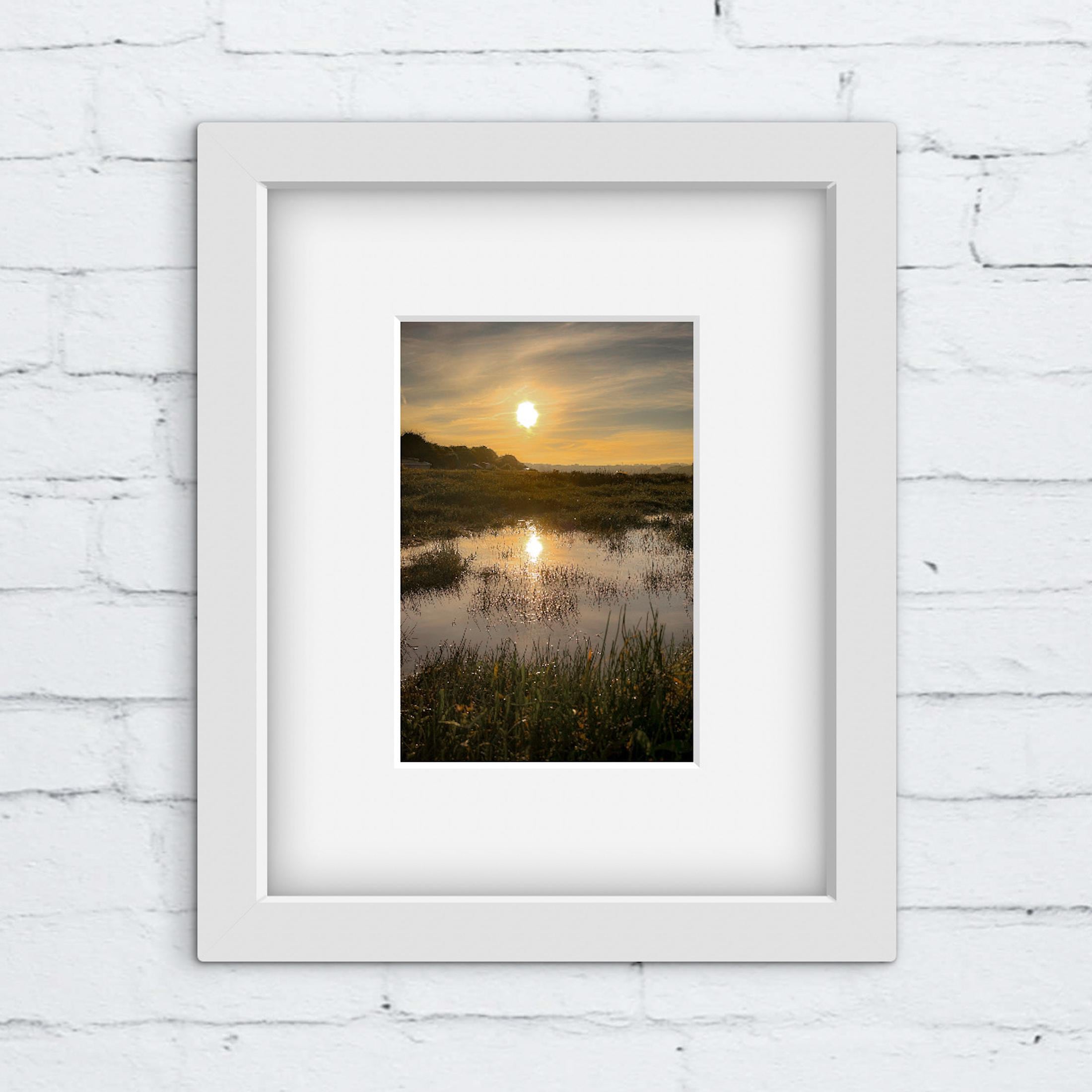 Framed photograph of a sunrise over a marshy field with a white brick wall background