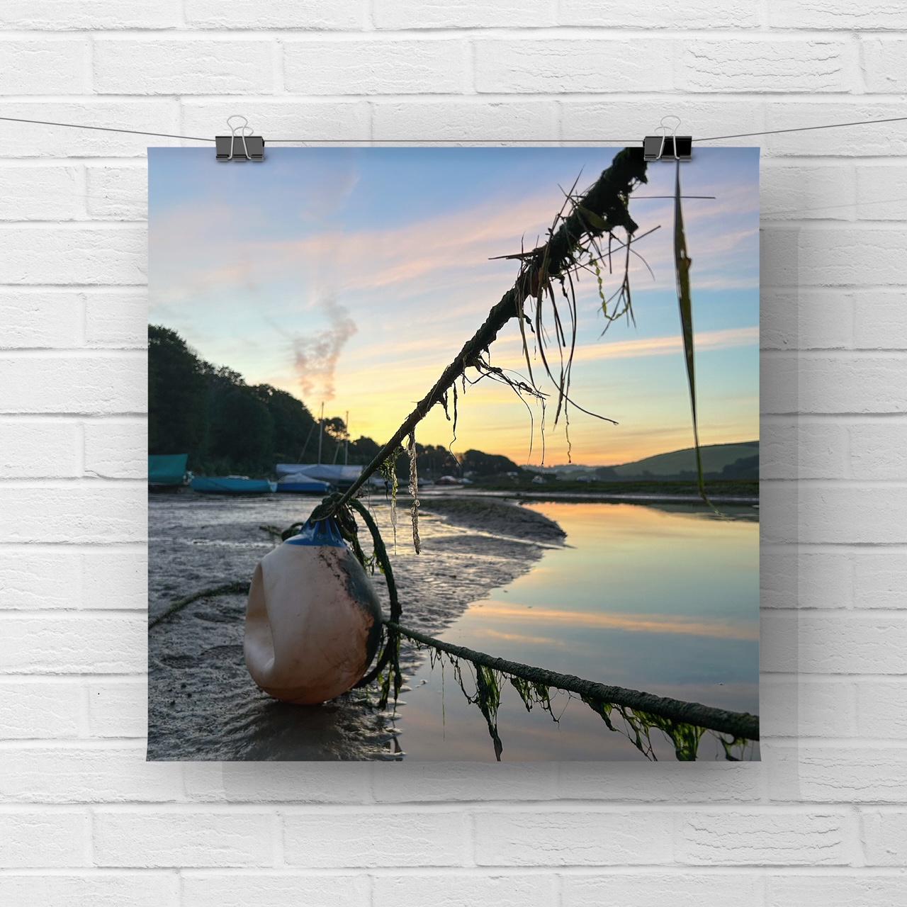 Poster of a sunriseover water with a buoy and mooring ropes,, pinned to a white brick wall.