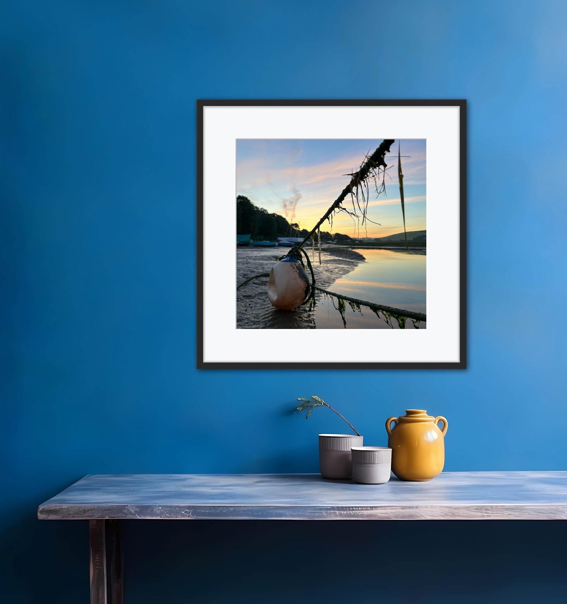 Framed photograph of a sunrise over water with moorings ropes and body on a blue wall above a wooden table with decorative items.