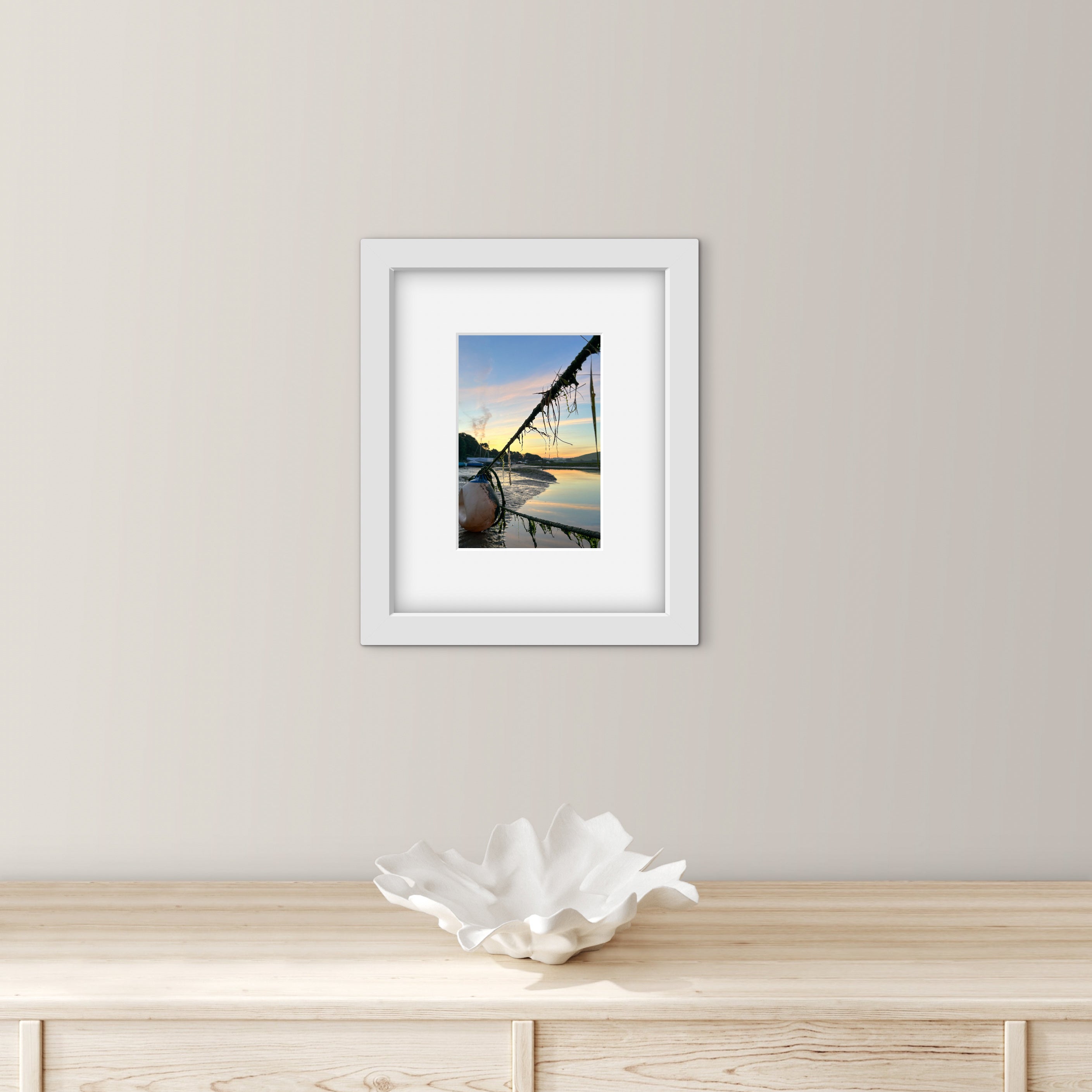 Framed artwork of a sunrise reflected over water with mooring ropes and a bouy on a wall above a wooden surface with a decorative bowl.