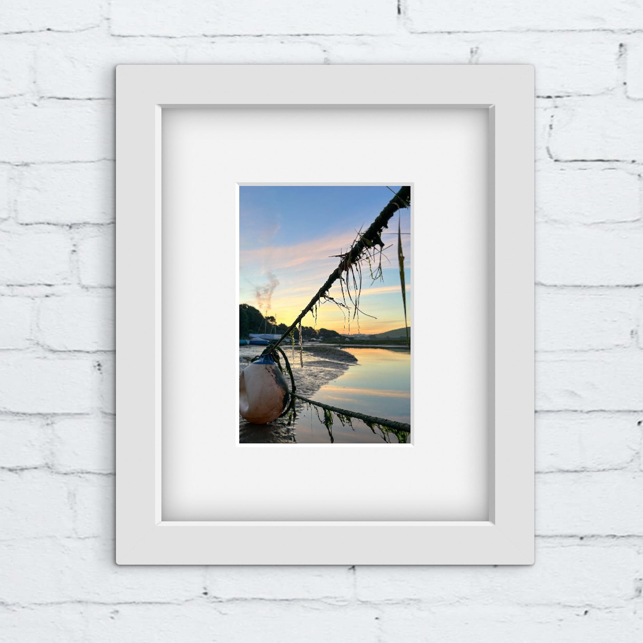 Framed photograph of a sunrise over water with mooring ropes on a river in a white frame on a white brick wall.
