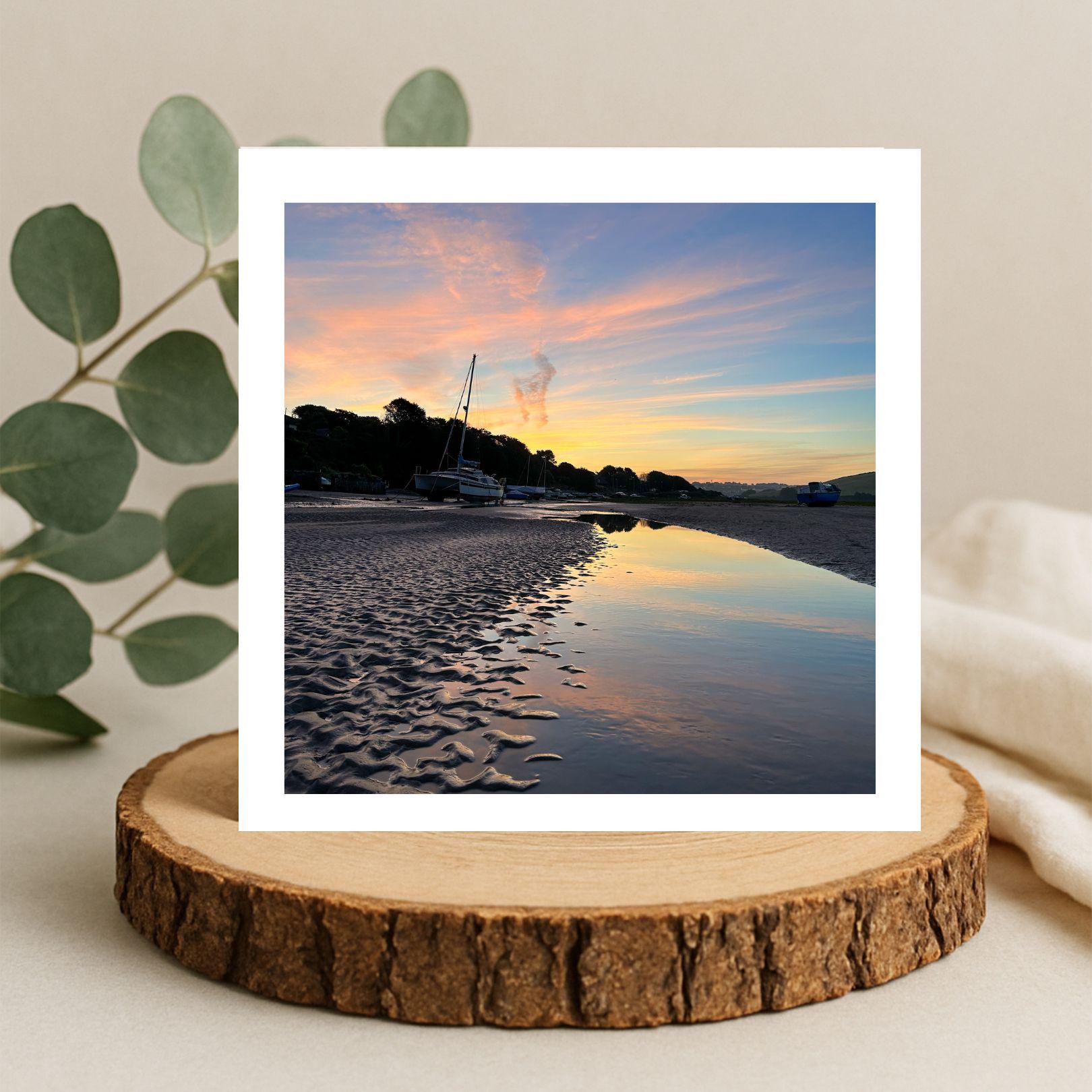 Greetings card of a sunset reflected  in water . Displayed on a wooden stand with a plant and blanket in the background.