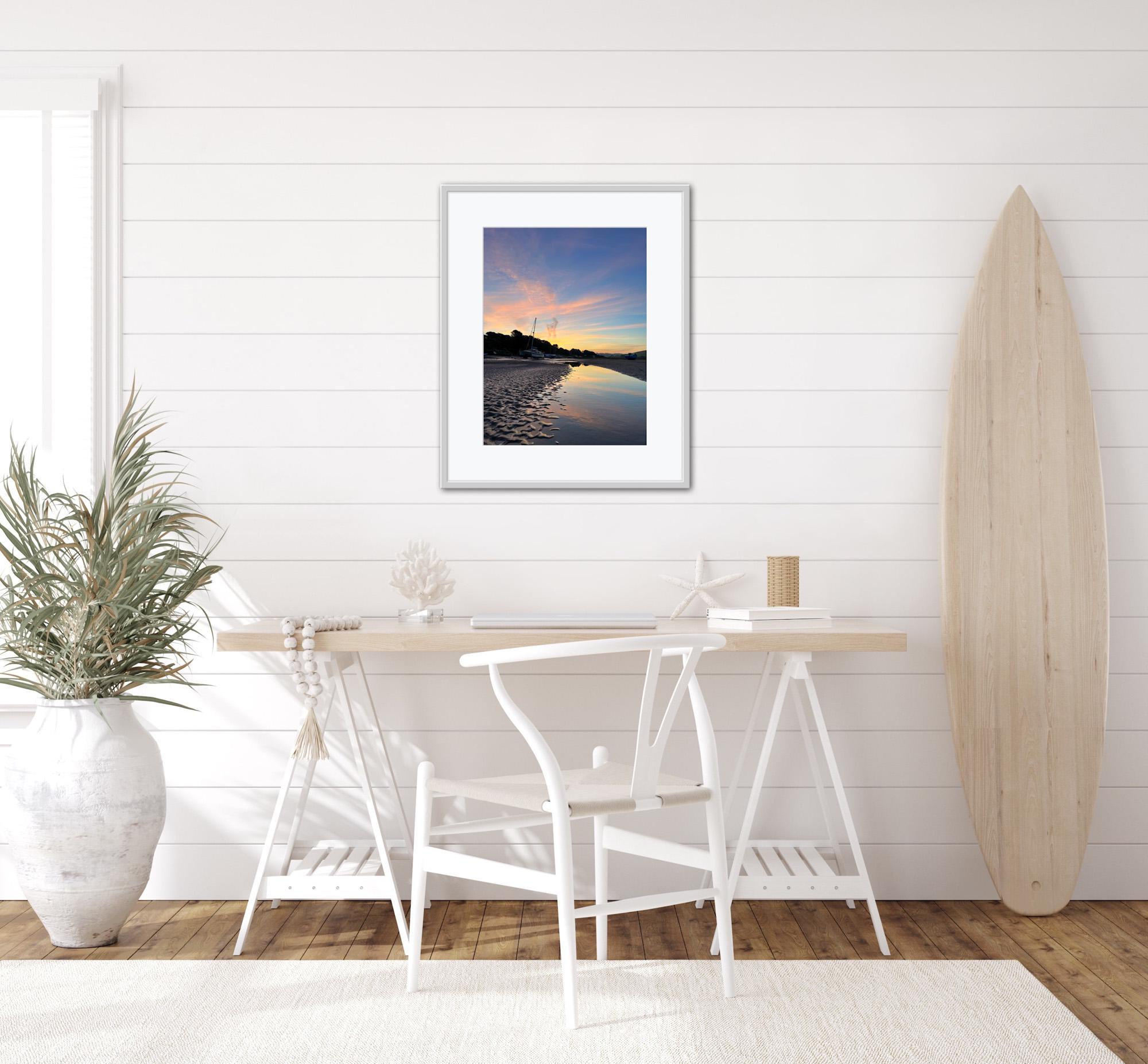 Modern interior with a framed beach sunrise reflected in water print, white chairs, and a surfboard against a white wall.