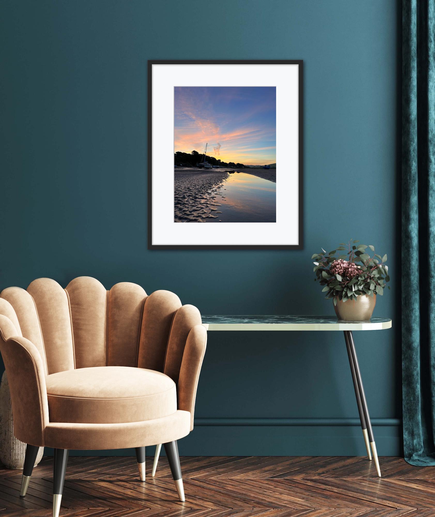Framed colourful sunrise artwork on a teal wall with a beige armchair and small table below.