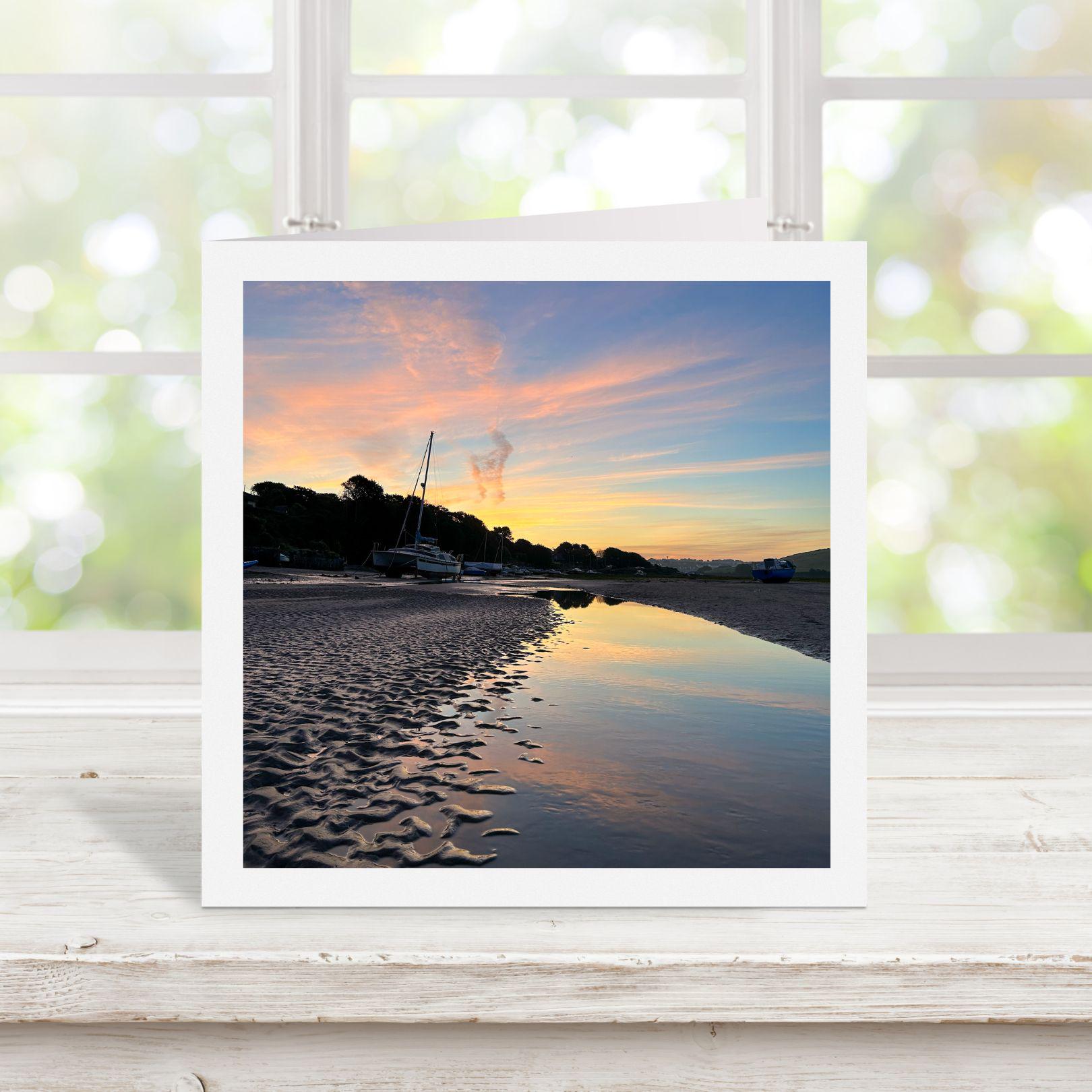 Square greetings card with photograph of a sunset over a beach with boats. Displayed on a wooden surface.