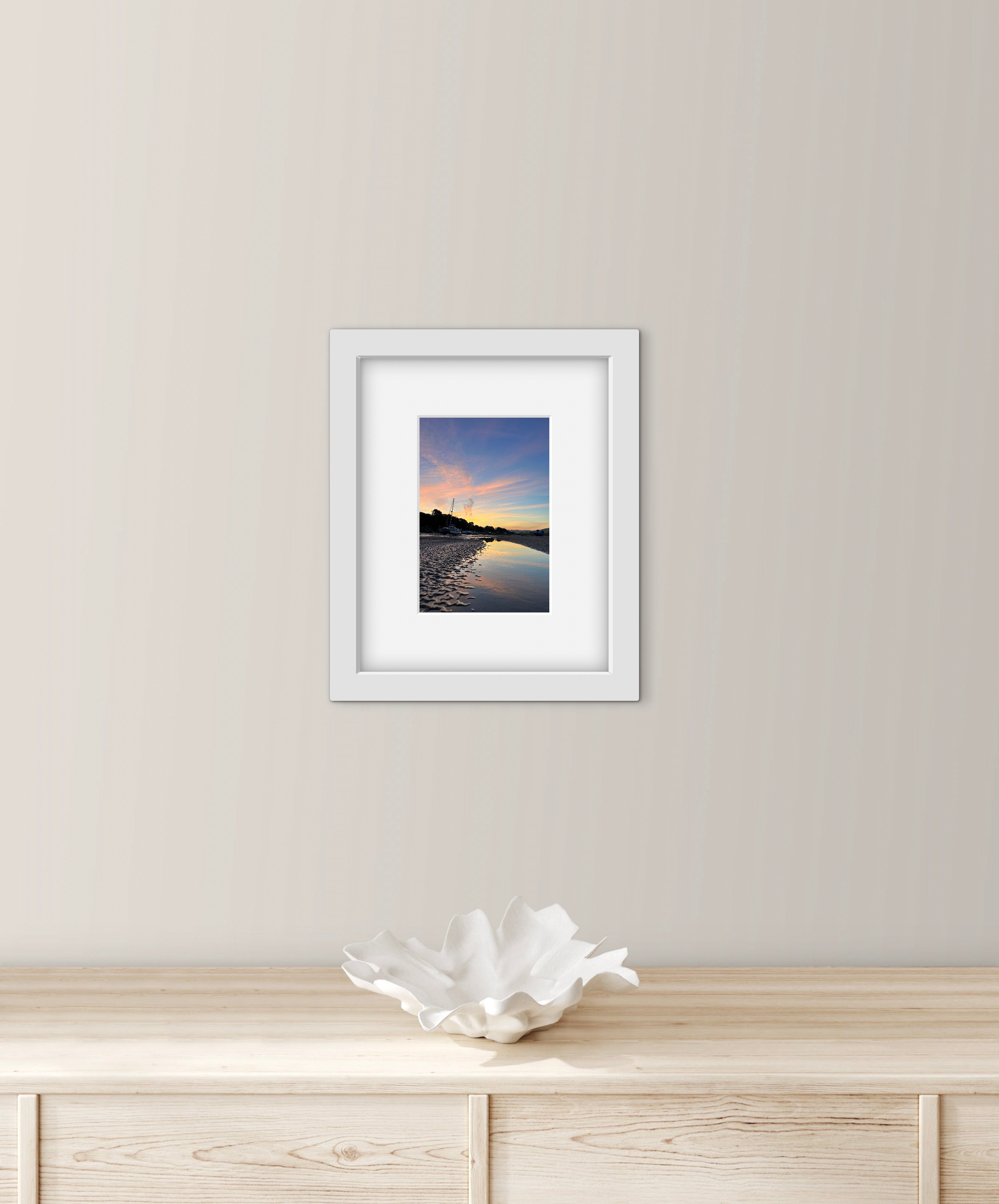 Framed photograph of a pastel sunset over water on a wall above a wooden cabinet with a decorative bowl.