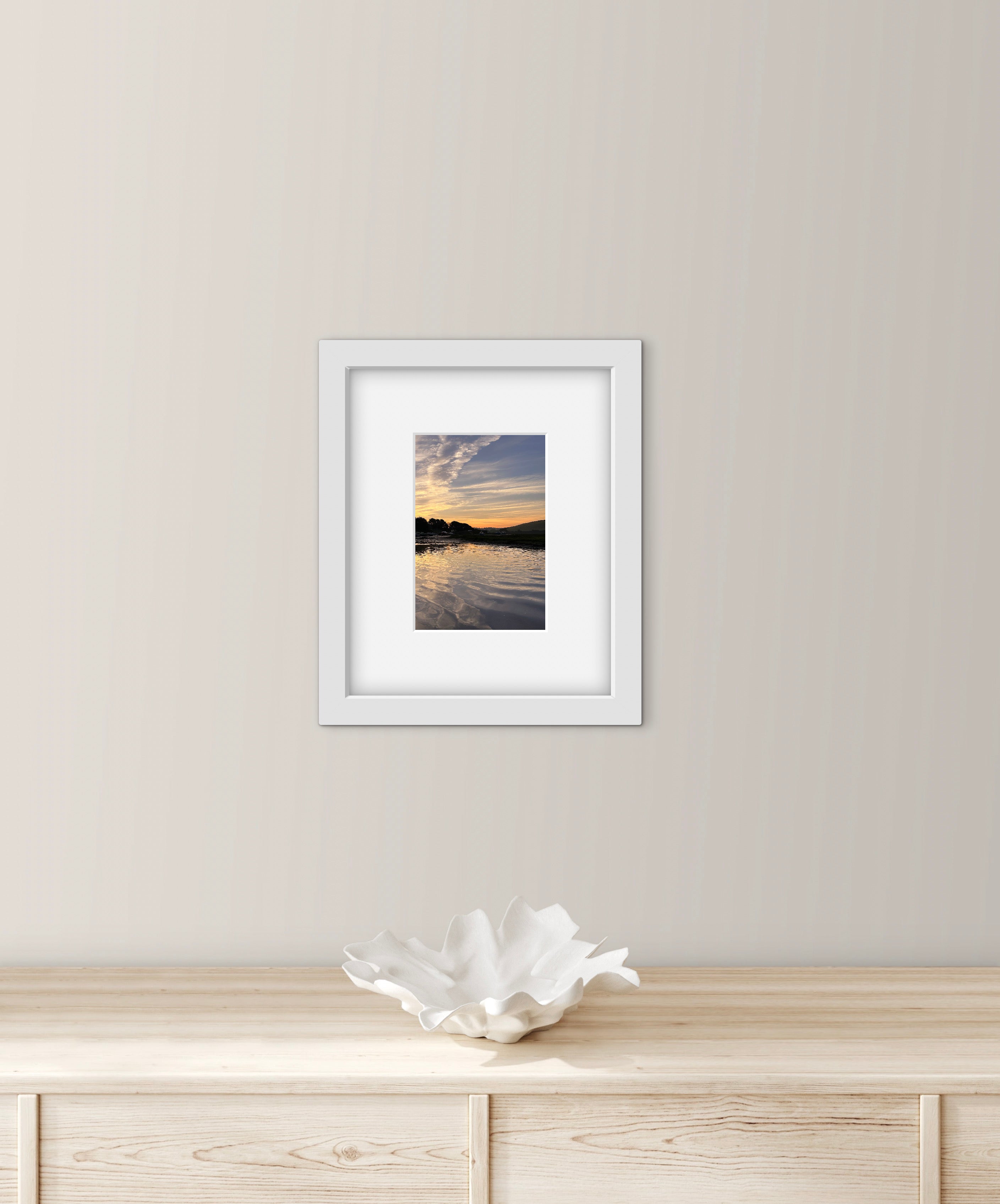 Framed photograph of a sunrise over water on a wall above a wooden cabinet with a decorative bowl.