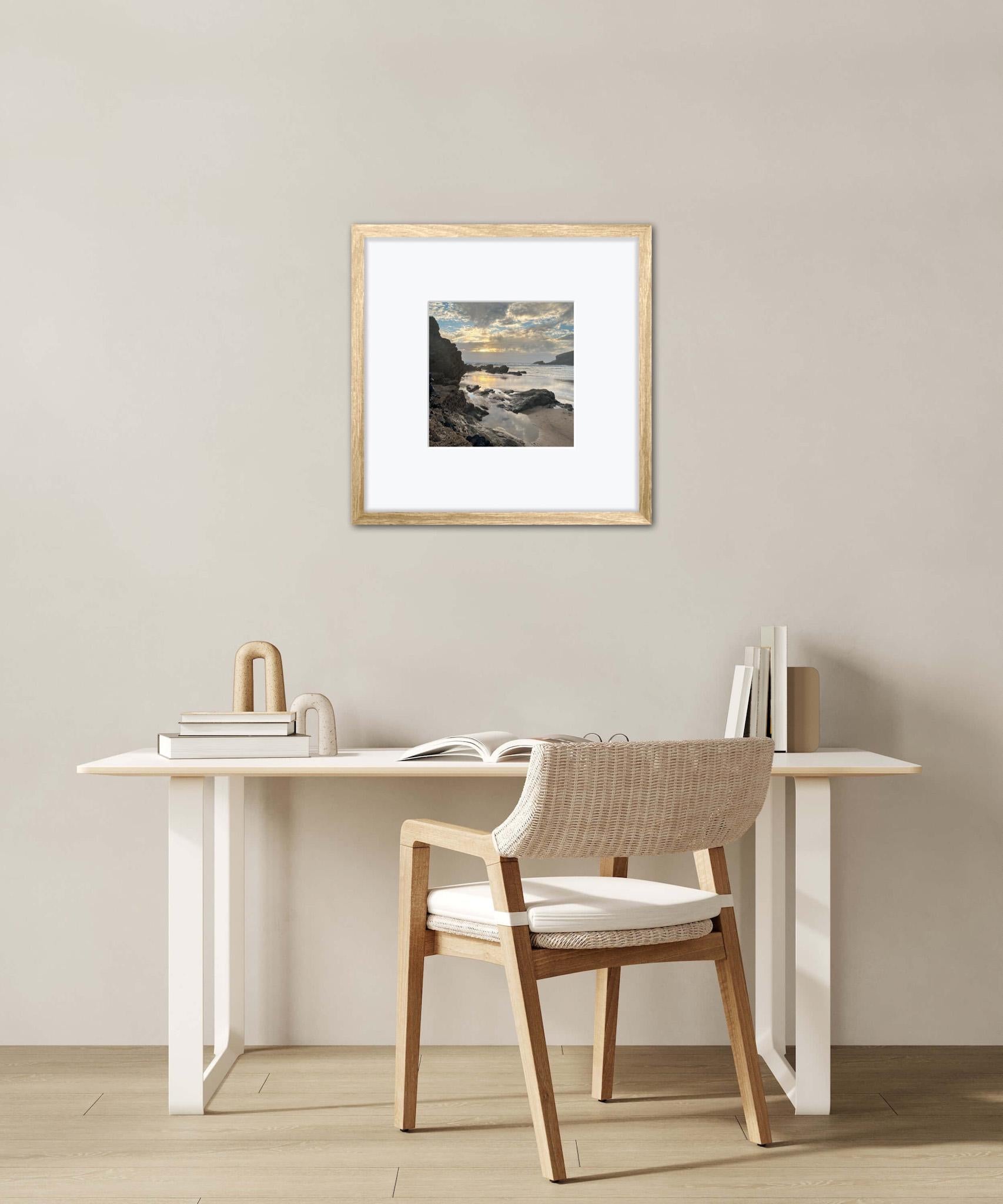 Minimalist interior with a desk, chair, and framed sunset over a rocky shore artwork on a plain wall.