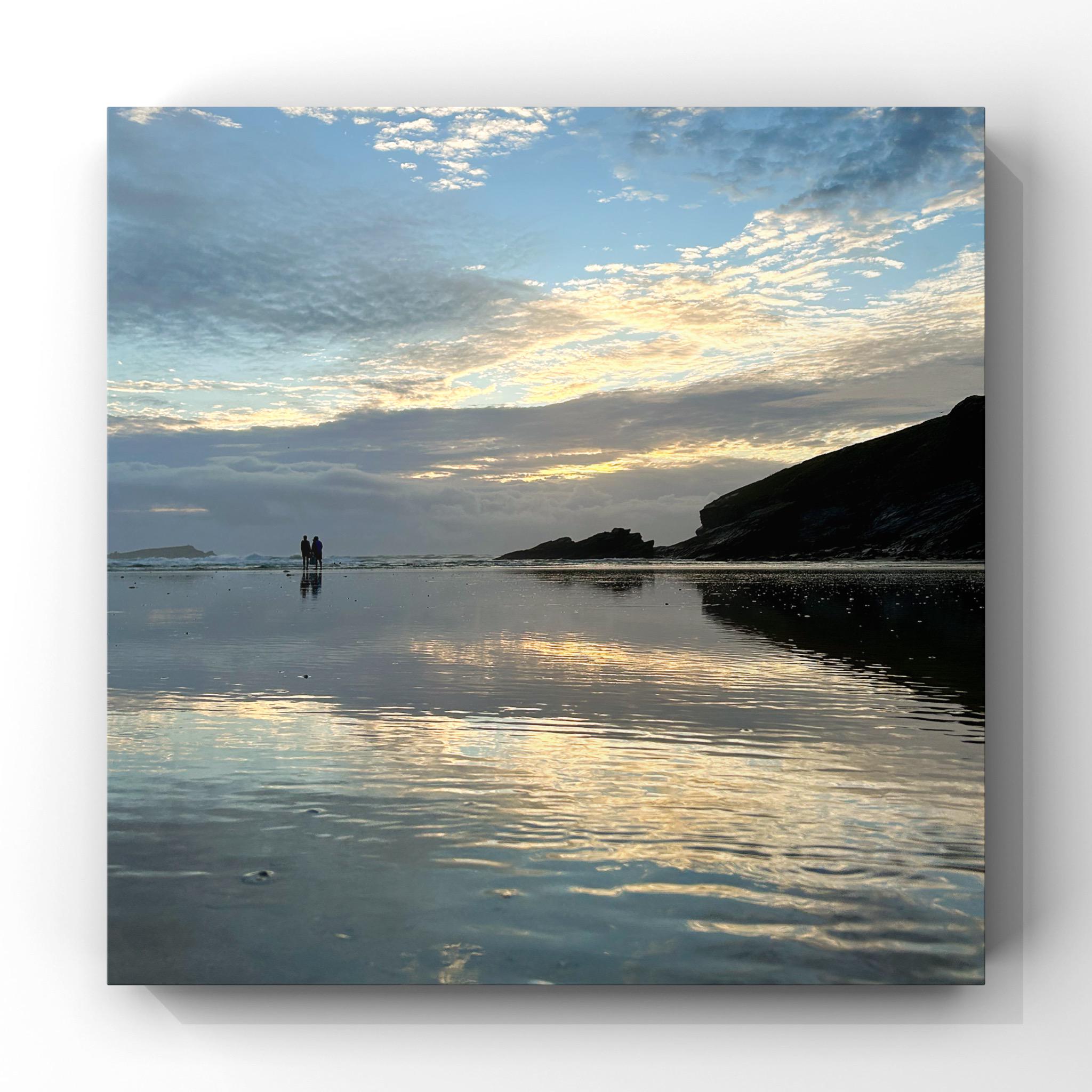 Two people standing on a beach at sunset with reflected clouds on the water.