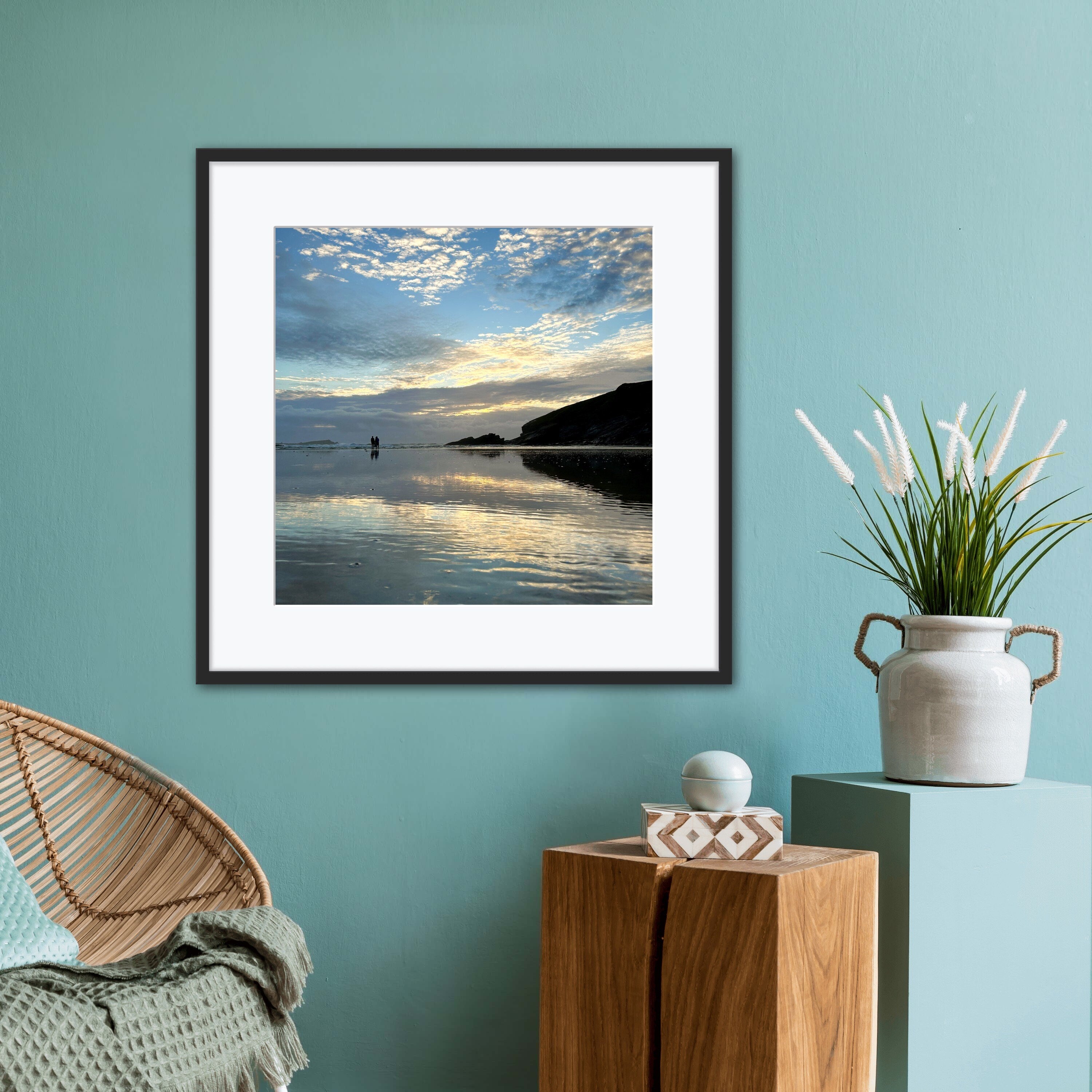 Framed artwork of a beach scene  at sunset reflected in wet sand on a teal wall with a chair and decorative items.
