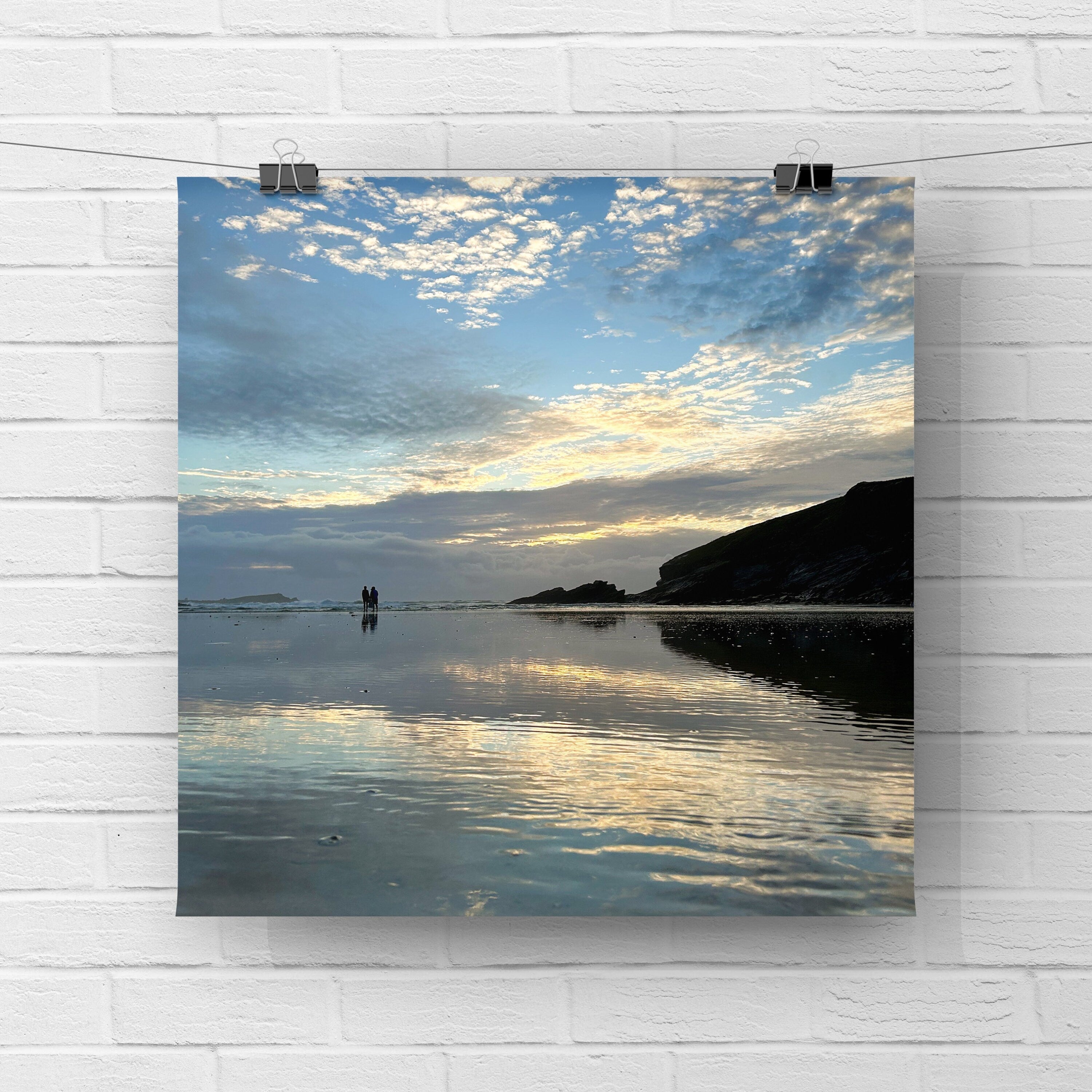 Poster of a beach scene with people standing on wet sand and a sunset reflection. The print is  hanging on a white brick wall.