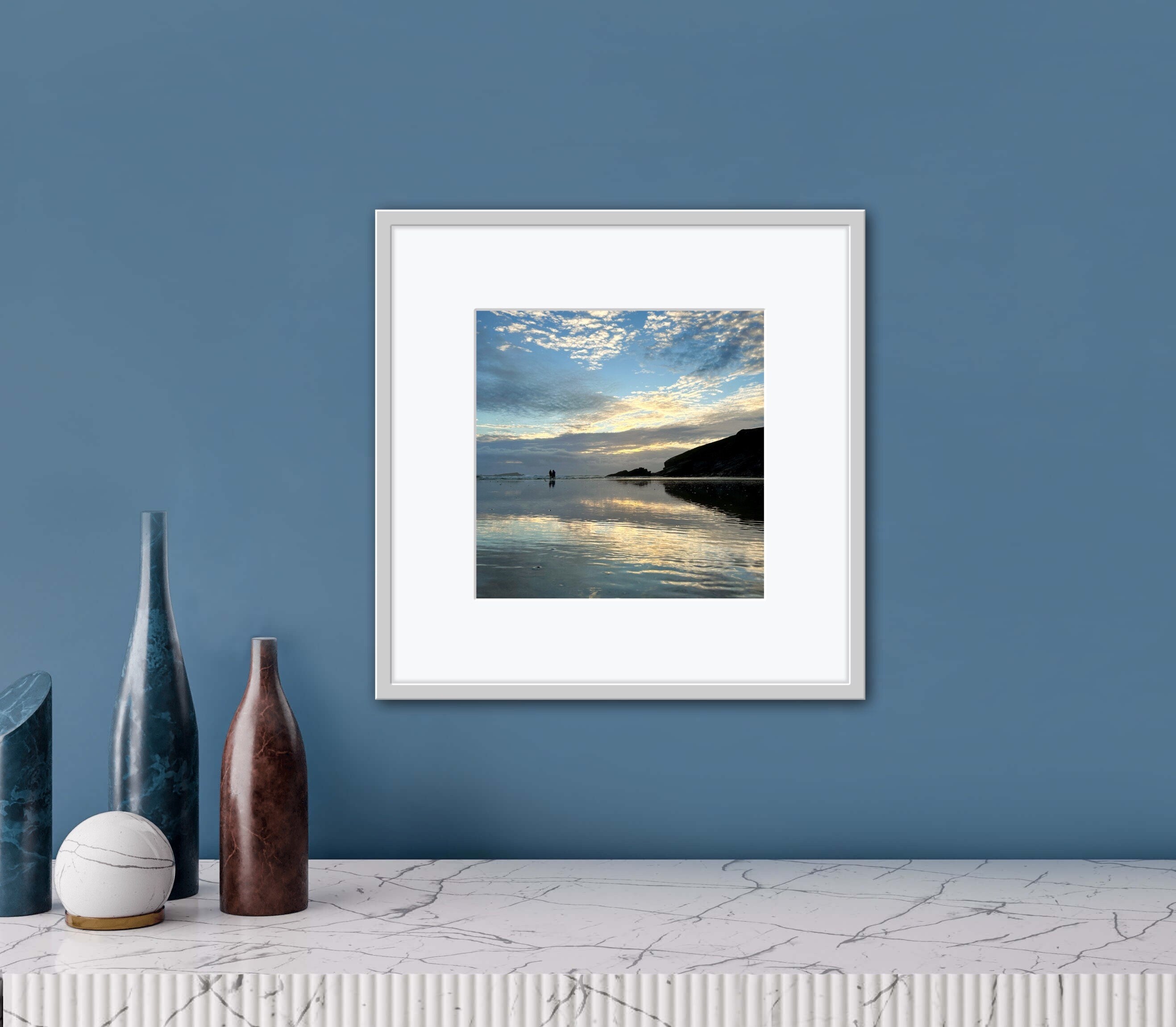 Framed artwork of a reflected sunset over water on a blue wall with decorative bottles below.