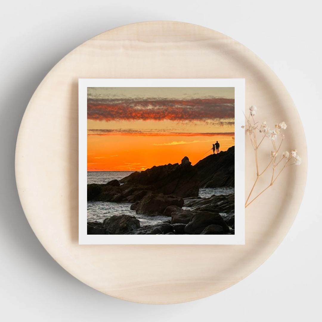 Greetings card of an sunset over a rocky landscape with two fishermen. Displayed on a beige plate.