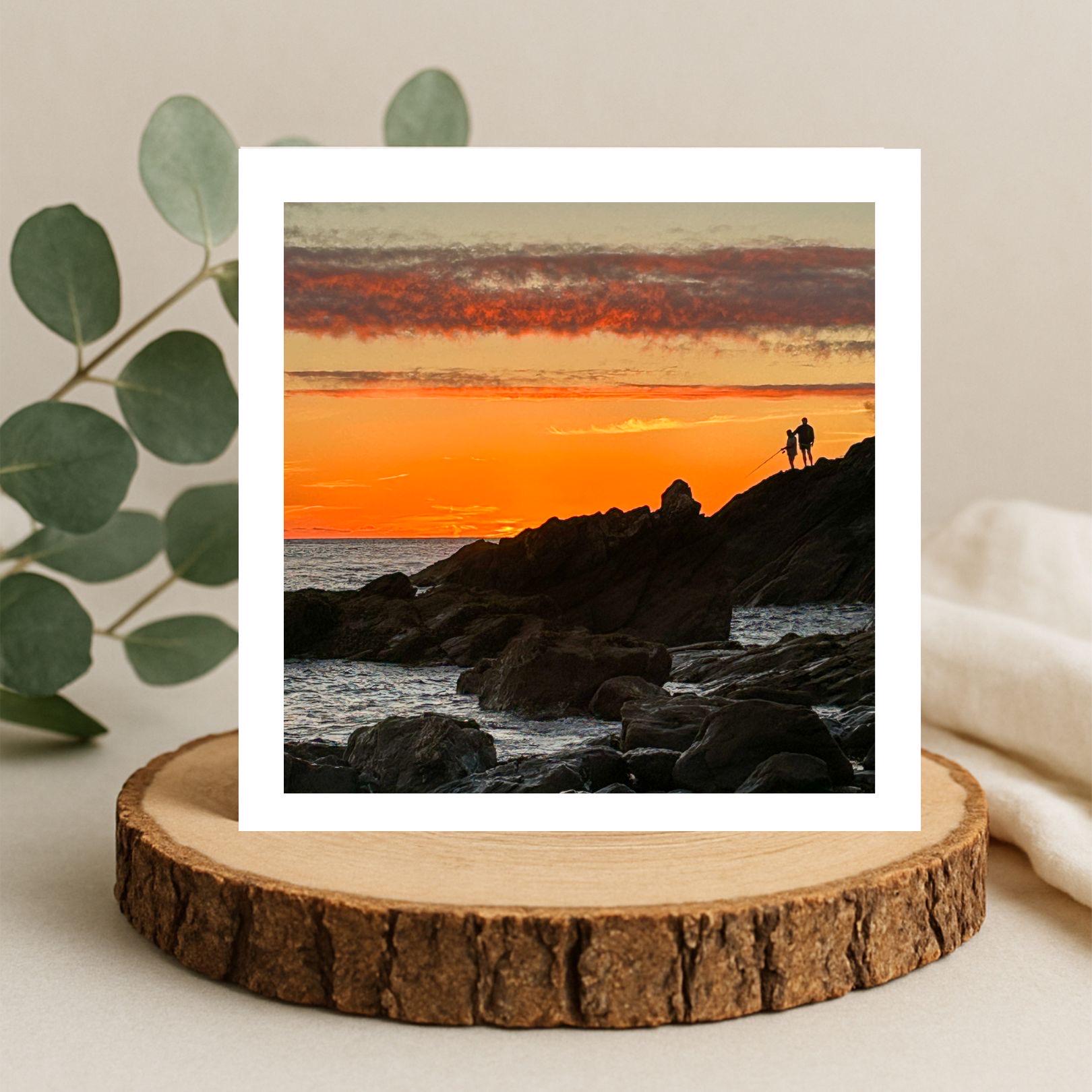 Sunset over a rocky landscape with two people standing on a rock fishing, greetings card framed by a white border on a wooden coaster.