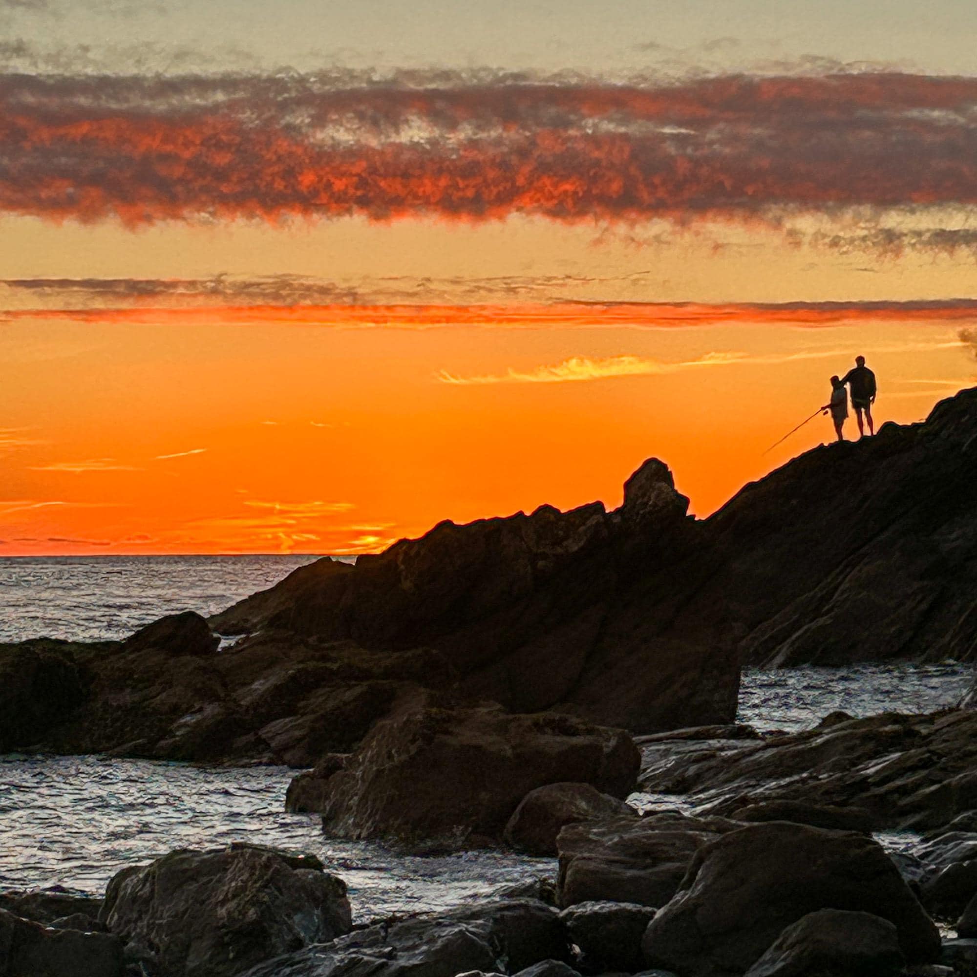 Two people standing on rocks fishing at sunset with a vibrant orange sky.