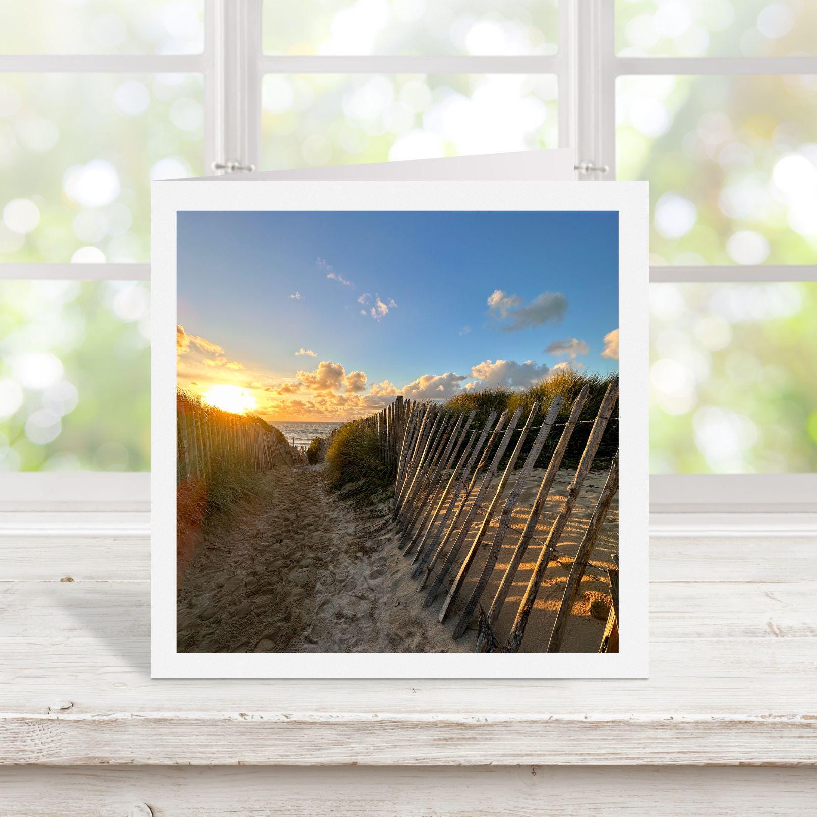 Square greetings card with photograph of a beach scene with a sunset over the ocean, displayed on a white surface.