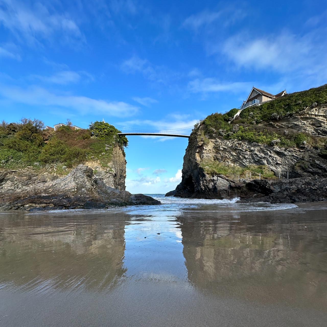 Bridge connecting two cliffs over a beach, reflected in wet sand, with clear blue sky