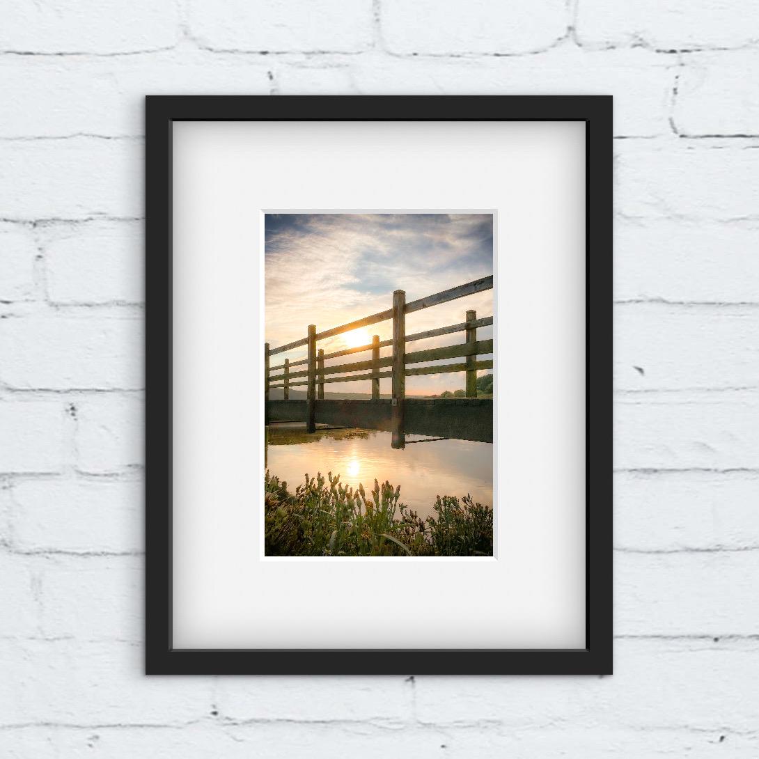 Framed photograph of a sunset over a wooden bridge and reflected in water. Black framed picture on a white brick wall.