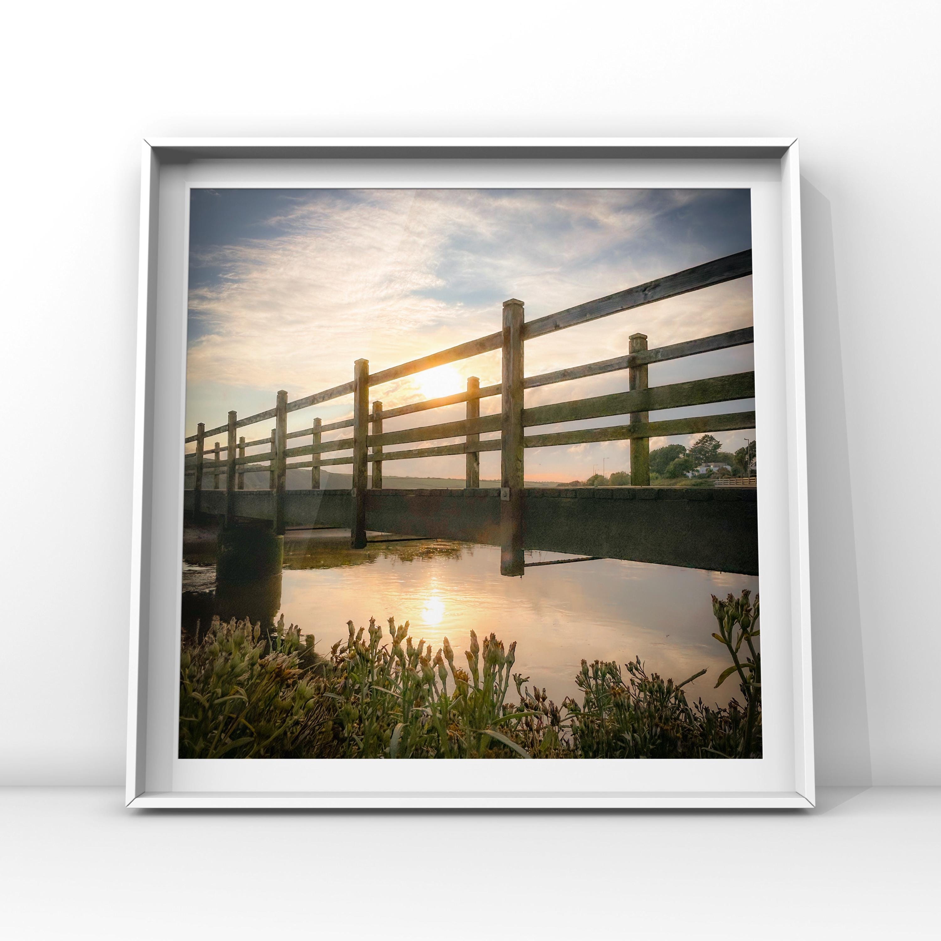 Framed photograph of a sunset over a wooden bridge reflected in water.
