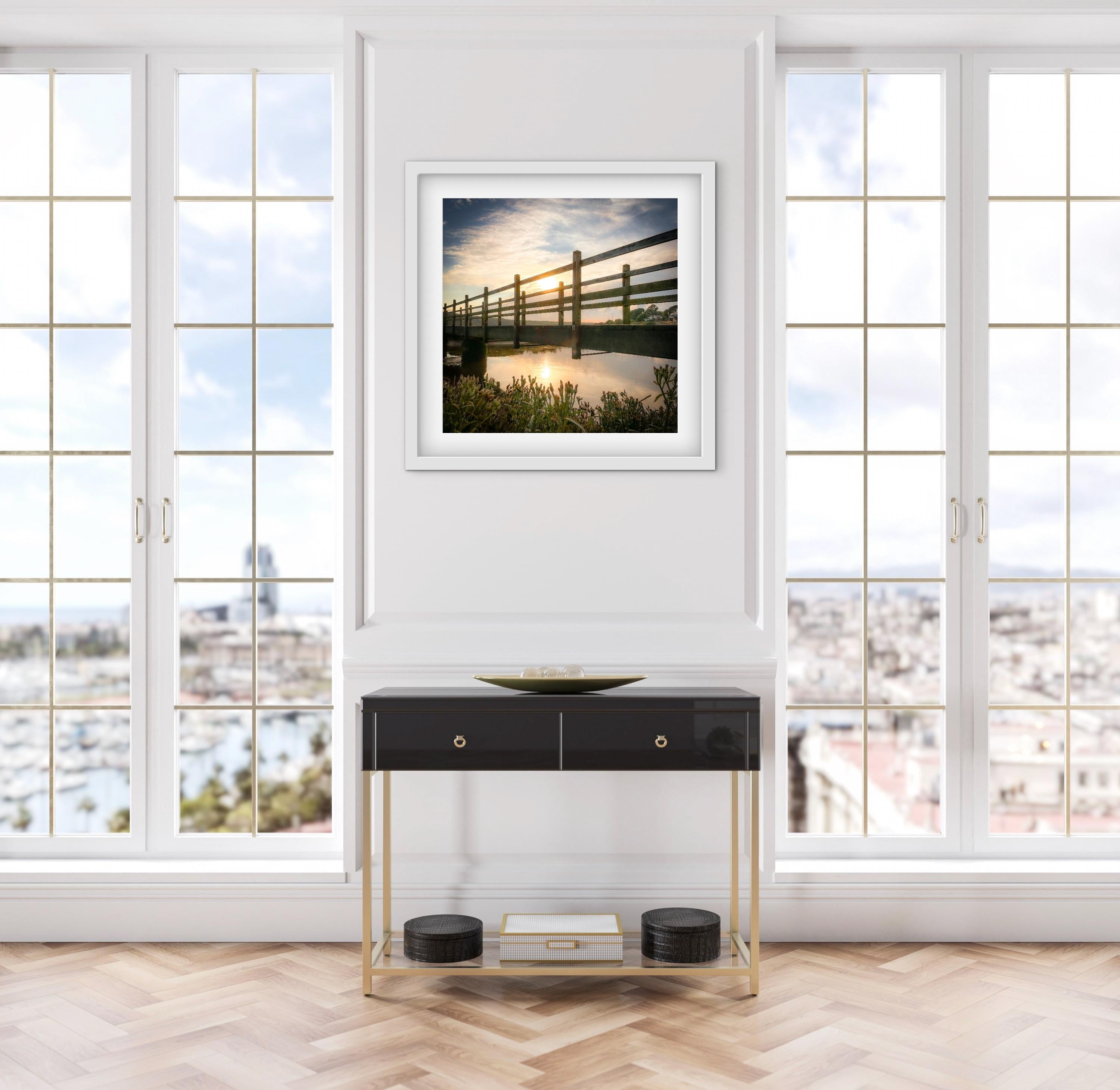 Modern console table with a framed picture above of a sunset behind a wooden bridge in a room with large windows.