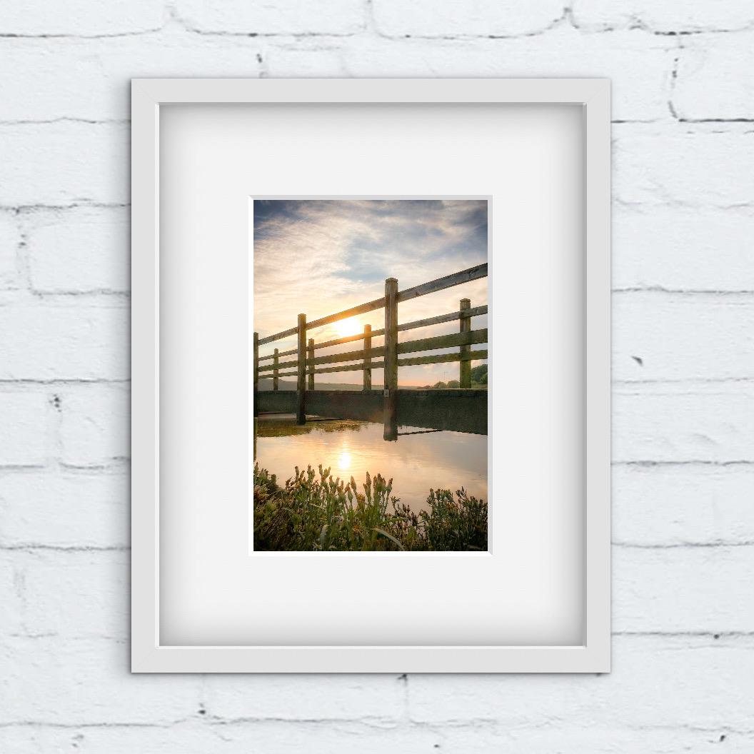 Framed photograph of a sunset over a wooden bridge over water. white framed photo on a white brick wall.