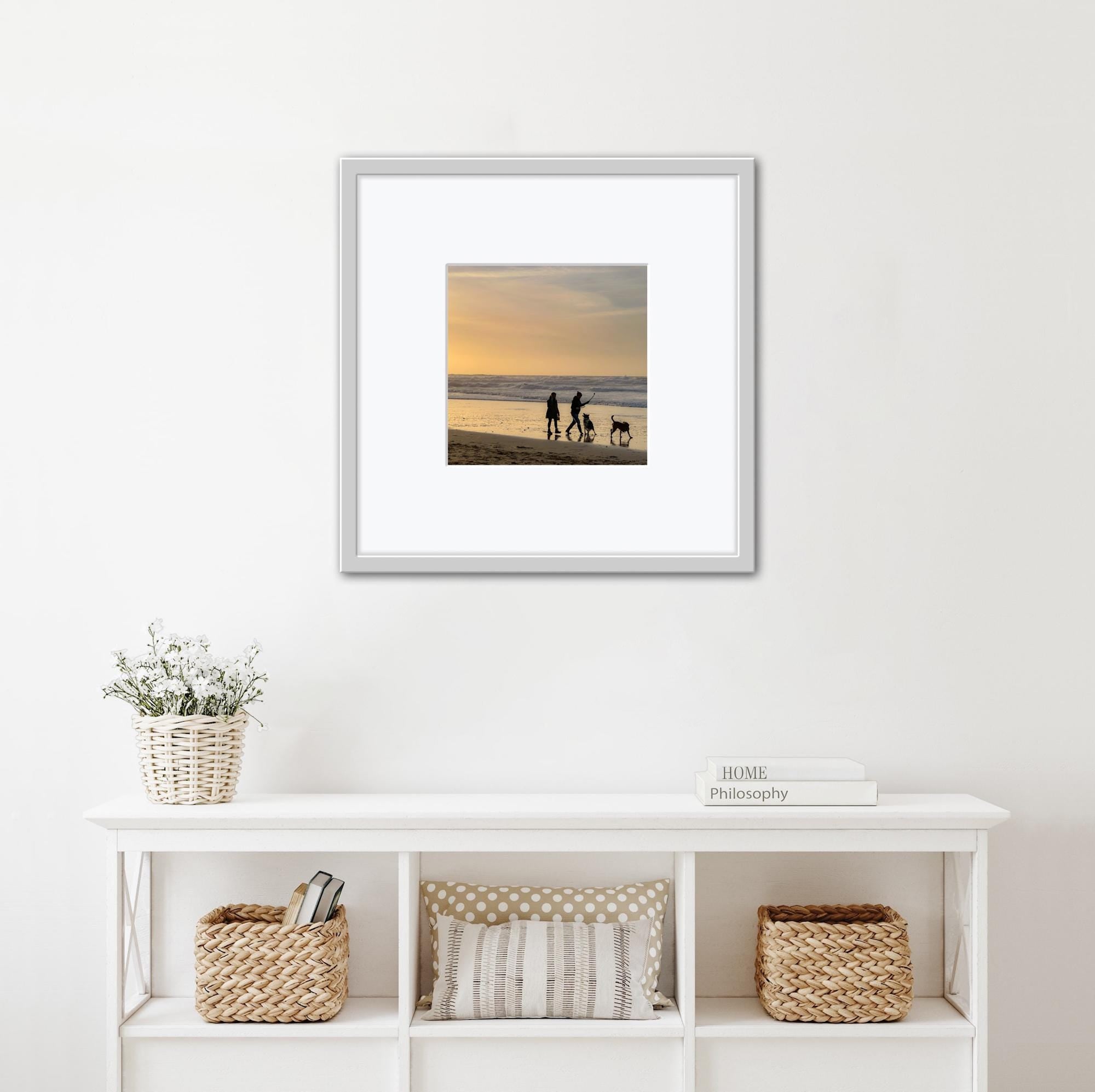 Framed photograph of a family with a dog on a beach at sunset, displayed on a wall above a white shelf.