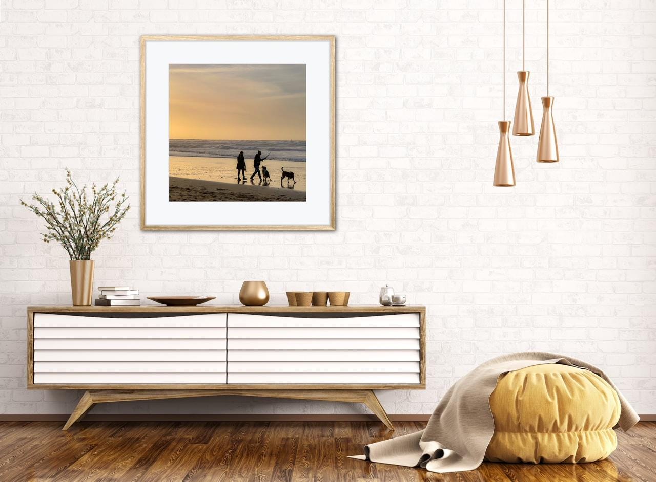 Modern living room with a framed picture of a beach scene and two people walking dogs at sunset, wooden console table, and decorative items.