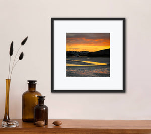 Framed artwork of an orange sunrise over water on a wall above a wooden shelf with decorative items.
