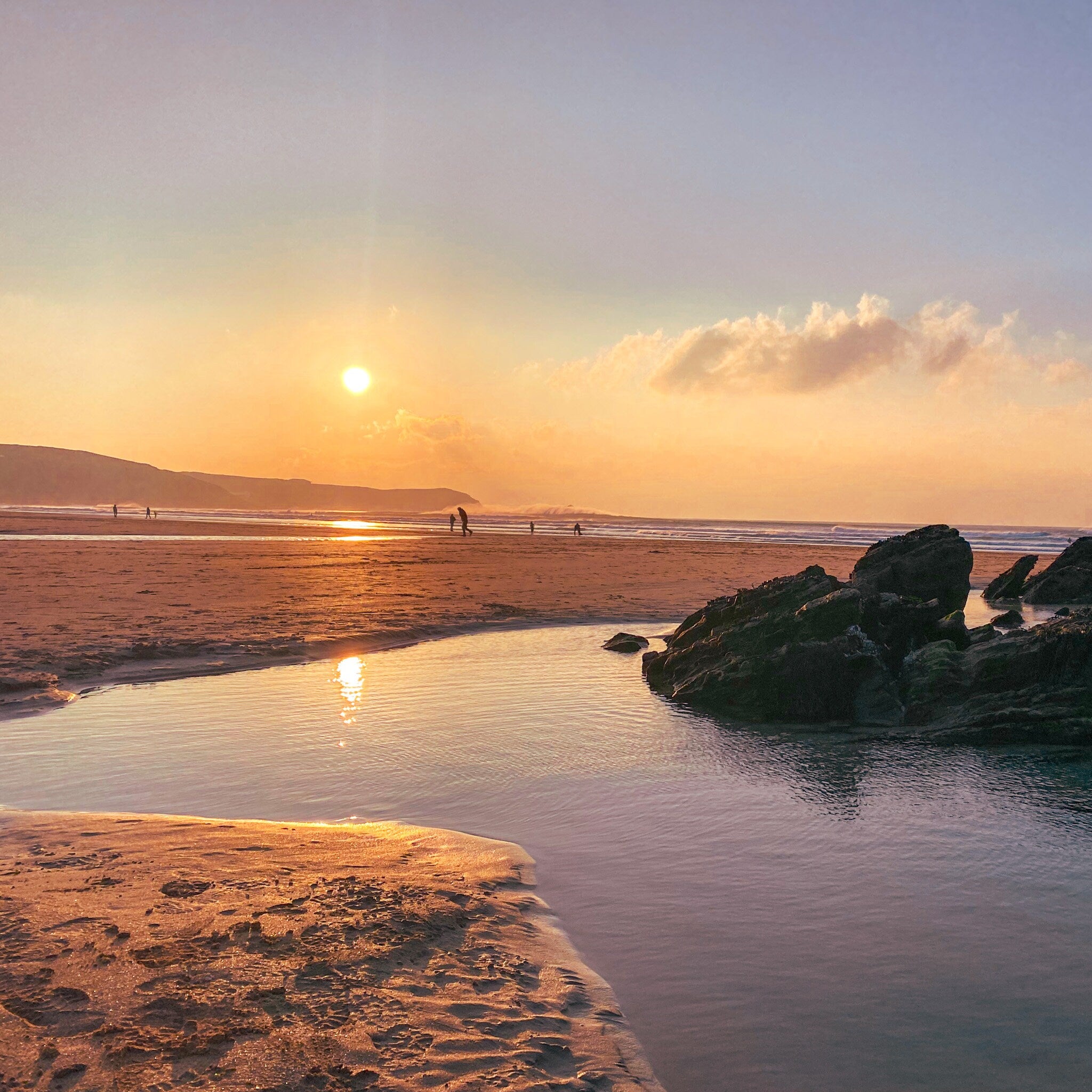 Sunset at a beach with people walking along the shore and large rocks  and rockpools in the foreground.
