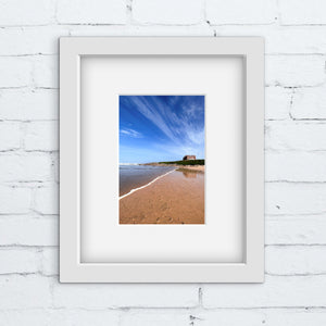 Framed photograph of a beach scene with an old hotel on a headland in a white frame on a white brick wall.