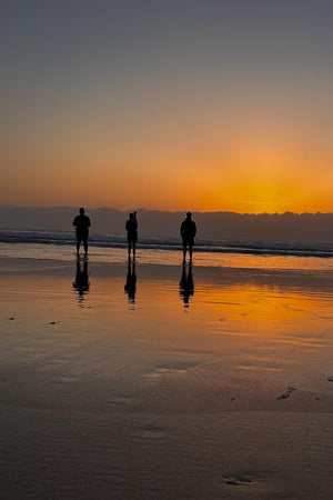 Three people standing on a beach at sunset, silhouetted against the orange sky.