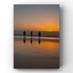 Three people standing on a beach at sunset, reflected in the wet sand.