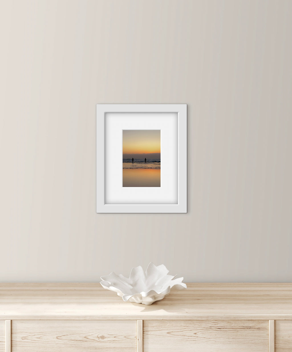 Framed artwork of two people going for a sea swim at sunset on a wall above a wooden surface with a decorative bowl.