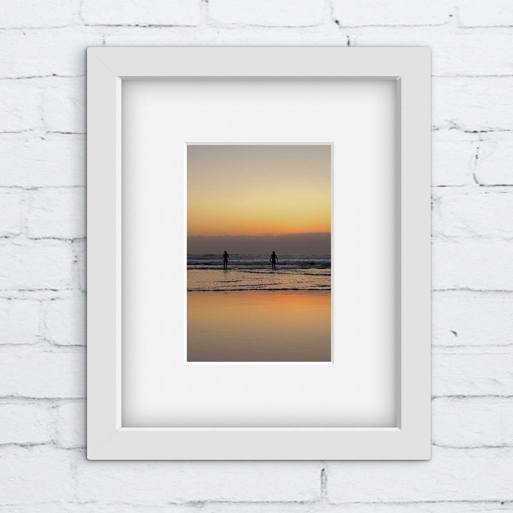 Framed artwork of a sunset over water with two silhouettes, on a white brick wall.