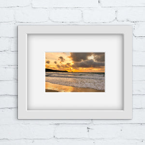 Framed photograph of a sunset over a beach on a white brick wall.