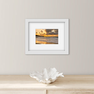 Framed artwork of a sunset over water on a wall above a wooden surface with a decorative shell.