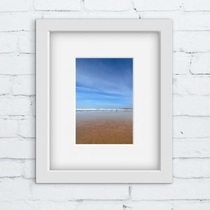 Framed photograph of a beach scene with blue sky and red sand on a white brick wall.